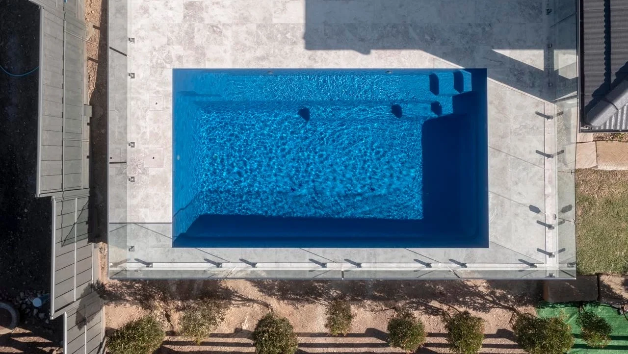 The Reflection 6.00m fibreglass swimming pool is installed in the heart of a hill house with white tiles and pool shades in the background. Its stunning Sapphire Blue colour stands out from the white house and surrounding mountainous greenery.