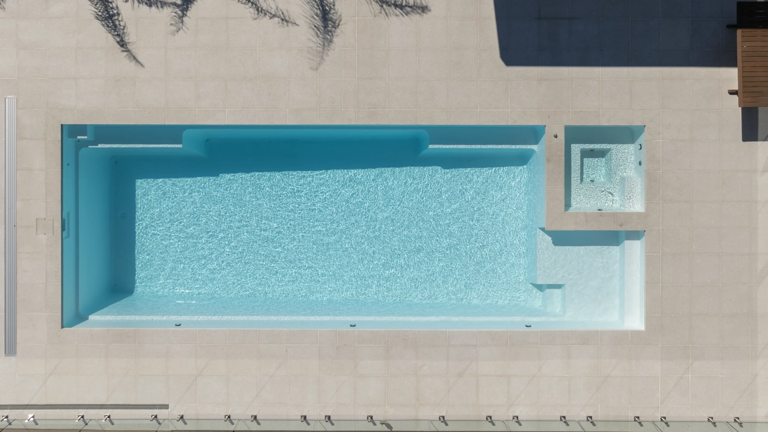 The Absolute 12.00m fibreglass swimming pool is in the middle of the backyard, with white tiles and greenery surrounding it. The pool colour is Shimmer White, which makes the fibreglass swimming pool stand out from the white modern house background and the morning blue sky.