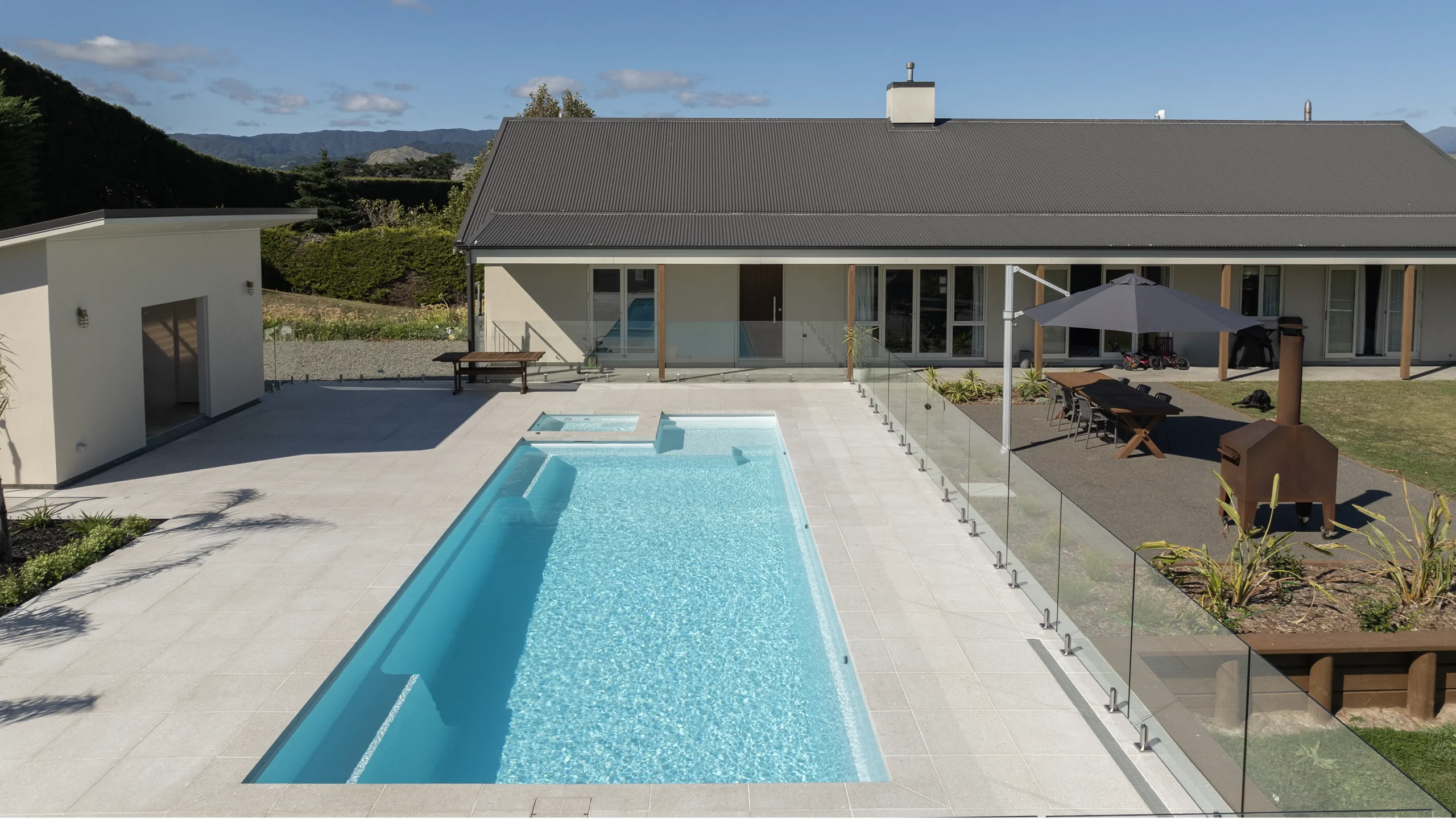 The Absolute 12.00m fibreglass swimming pool is in the middle of the backyard, with white tiles and greenery surrounding it. The pool colour is Shimmer White, which makes the fibreglass swimming pool stand out from the white modern house background and the morning blue sky.