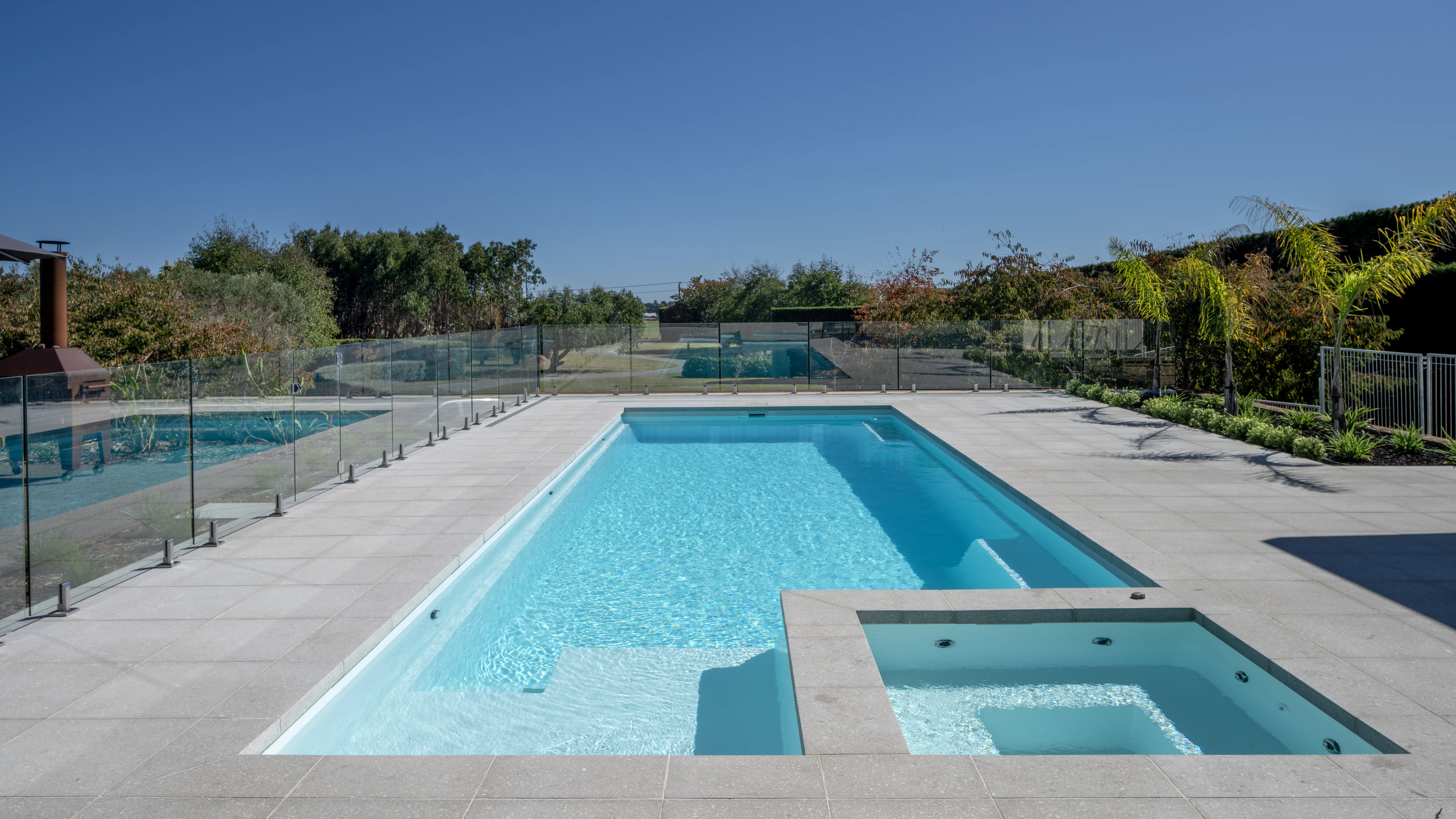 The Absolute 12.00m fibreglass swimming pool is in the middle of the backyard, with white tiles and greenery surrounding it. The pool colour is Shimmer White, which makes the fibreglass swimming pool stand out from the white modern house background and the morning blue sky.