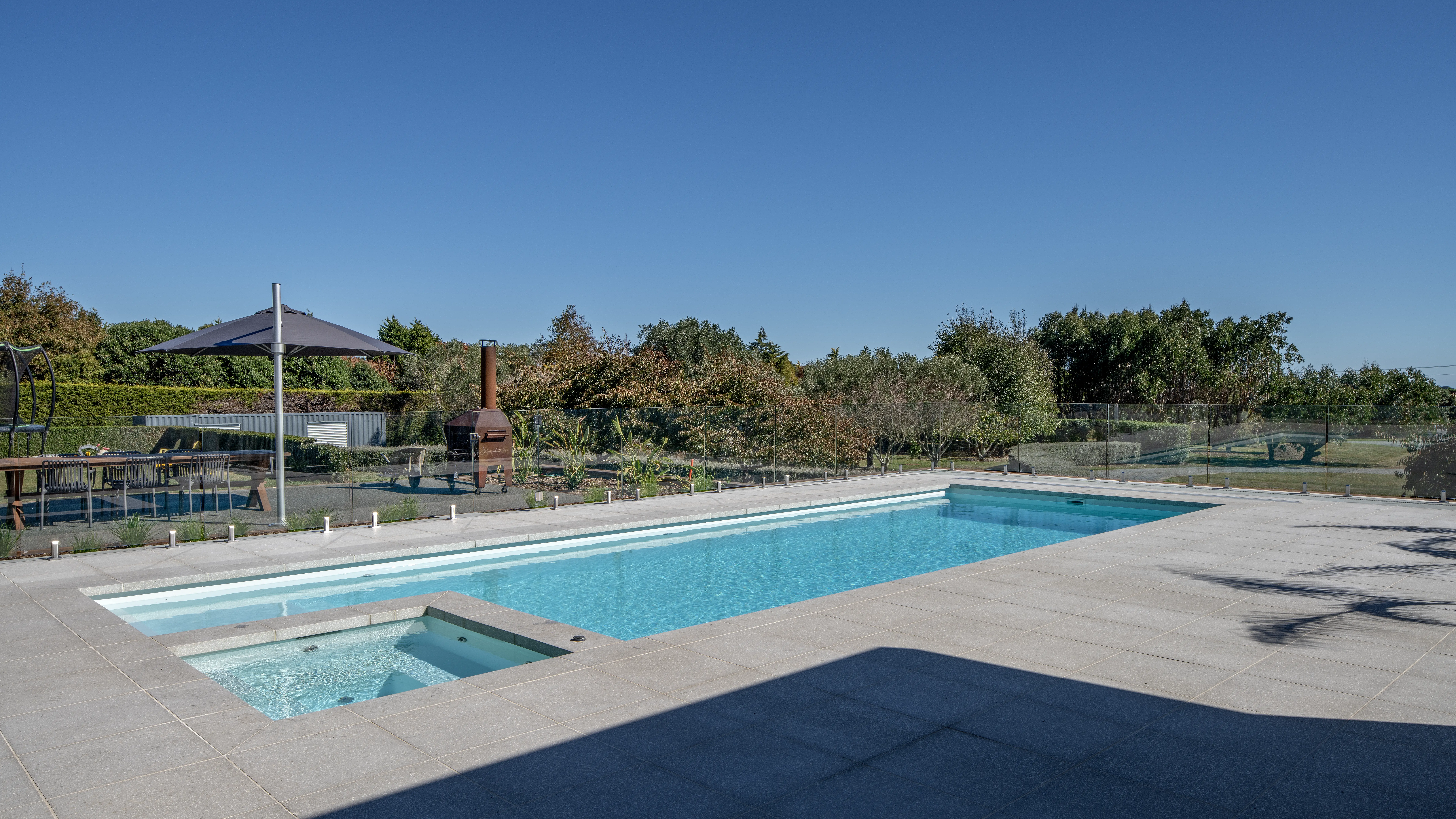The Absolute 12.00m fibreglass swimming pool is in the middle of the backyard, with white tiles and greenery surrounding it. The pool colour is Shimmer White, which makes the fibreglass swimming pool stand out from the white modern house background and the morning blue sky.