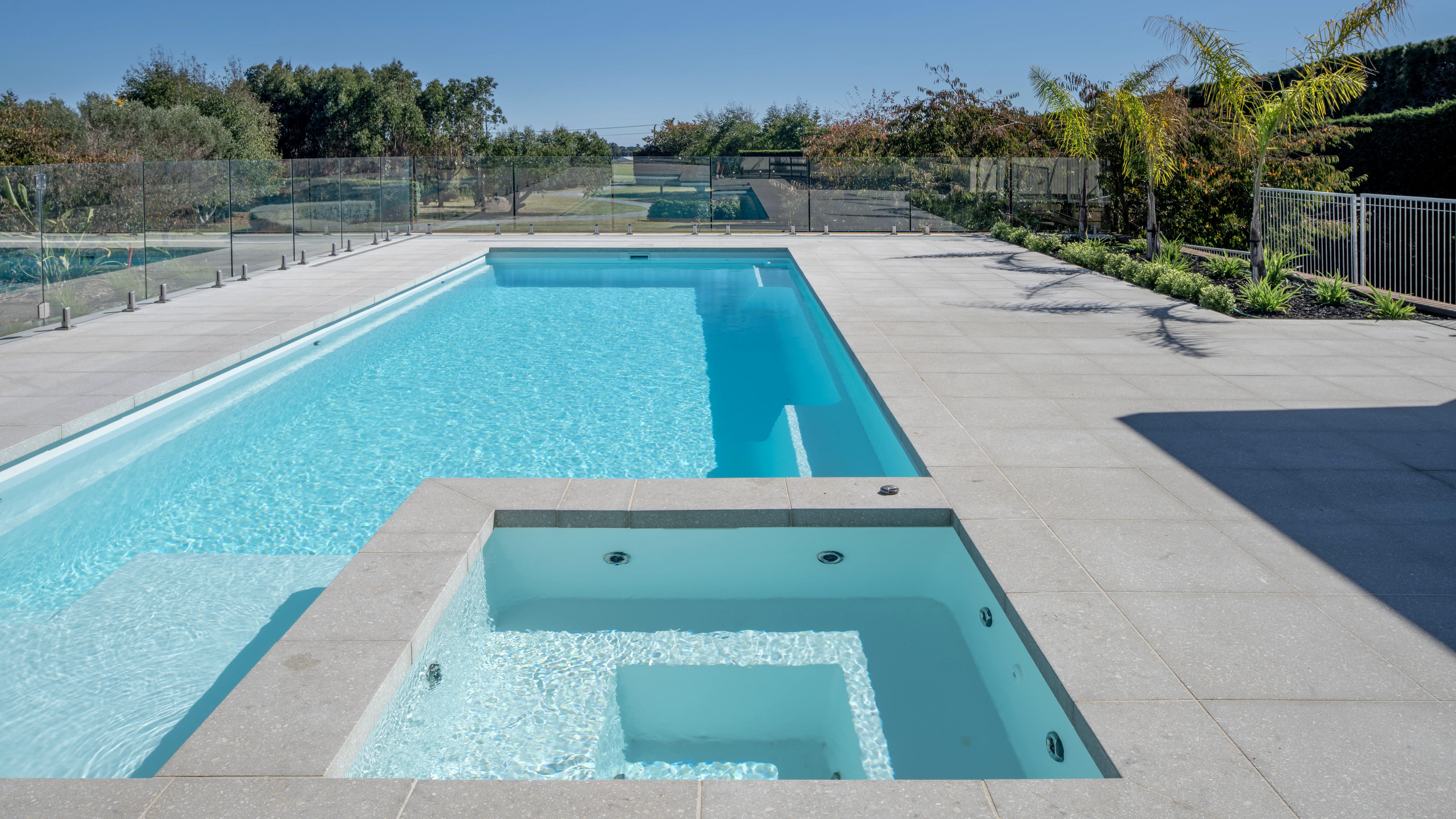 The Absolute 12.00m fibreglass swimming pool is in the middle of the backyard, with white tiles and greenery surrounding it. The pool colour is Shimmer White, which makes the fibreglass swimming pool stand out from the white modern house background and the morning blue sky.