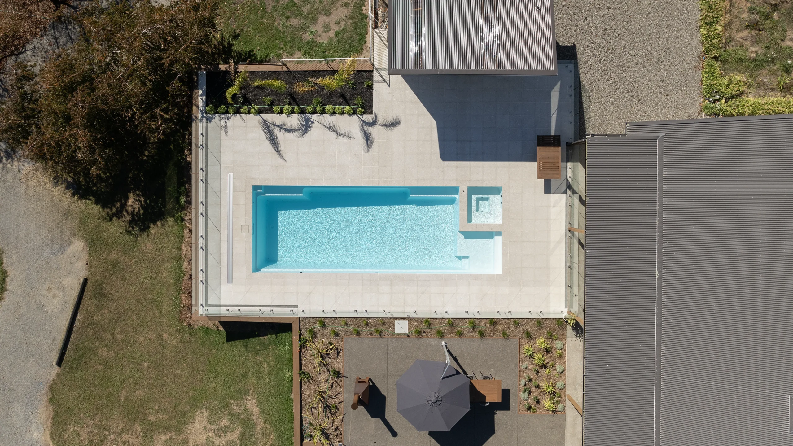The Absolute 12.00m fibreglass swimming pool is in the middle of the backyard, with white tiles and greenery surrounding it. The pool colour is Shimmer White, which makes the fibreglass swimming pool stand out from the white modern house background and the morning blue sky.