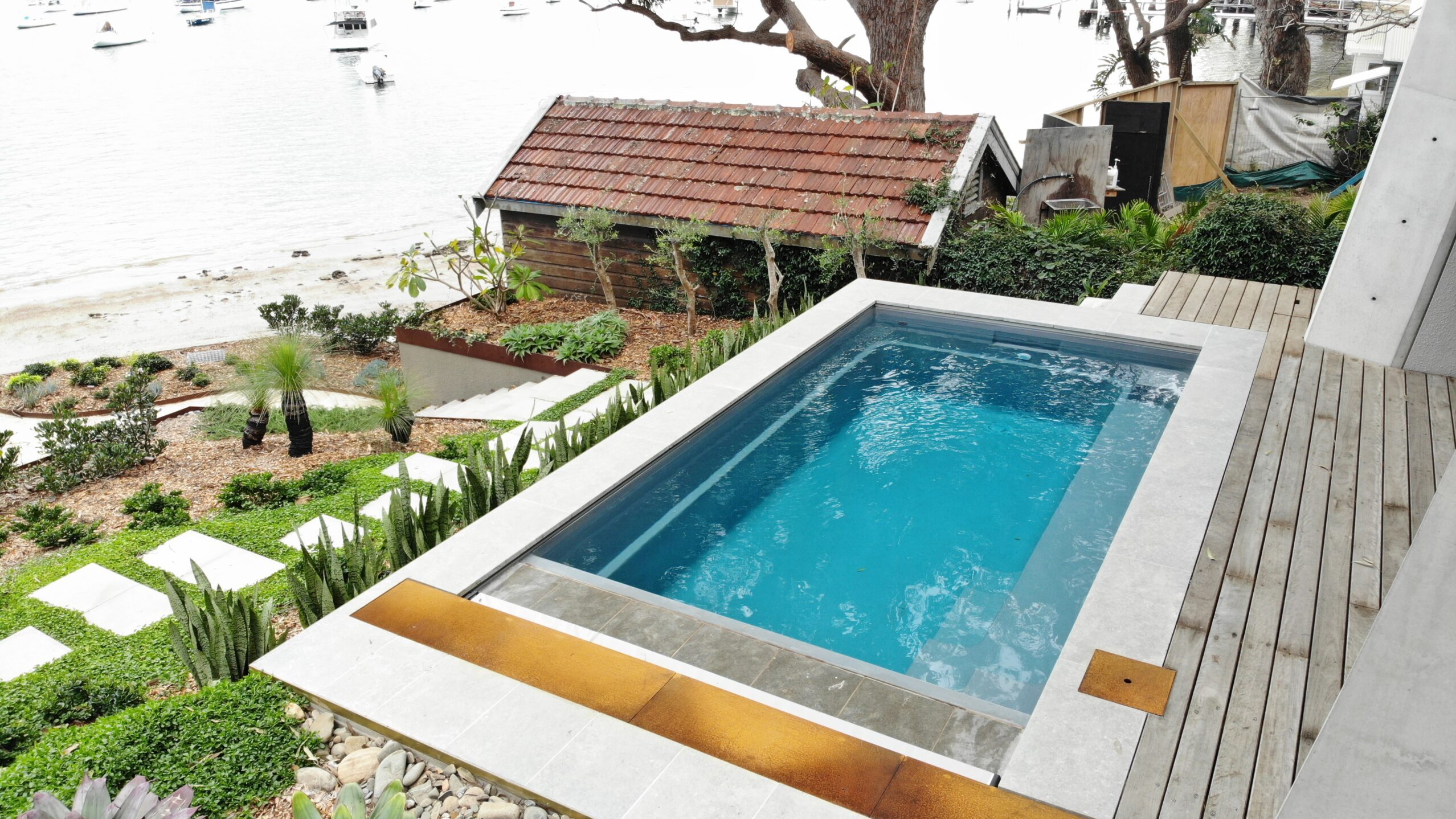 The Harmony 5.00m fibreglass swimming pool is installed in the wooden front yard of a beach house, with greenery and extra pool features surrounding it. The beautiful Graphite Grey colour stands out from the white-themed house, accented by extra yellow and grey detailing.
