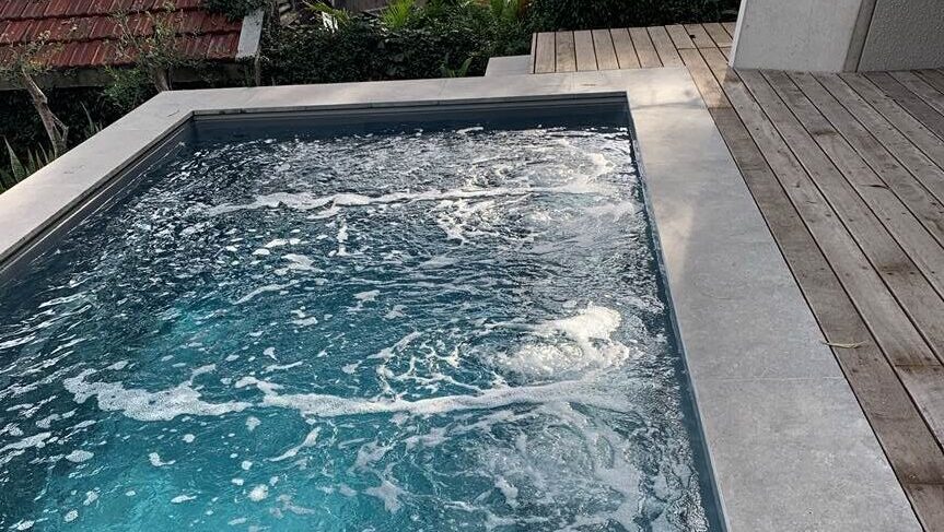 The Harmony 5.00m fibreglass swimming pool is installed in the wooden front yard of a beach house, with greenery and extra pool features surrounding it. The beautiful Graphite Grey colour stands out from the white-themed house, accented by extra yellow and grey detailing.