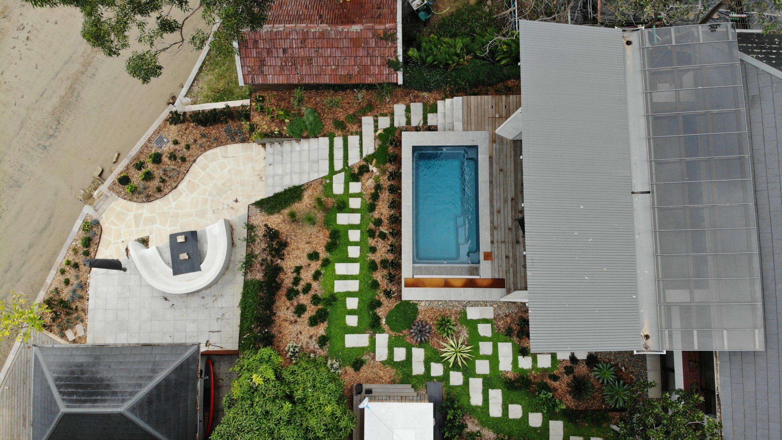 The Harmony 5.00m fibreglass swimming pool is installed in the wooden front yard of a beach house, with greenery and extra pool features surrounding it. The beautiful Graphite Grey colour stands out from the white-themed house, accented by extra yellow and grey detailing.