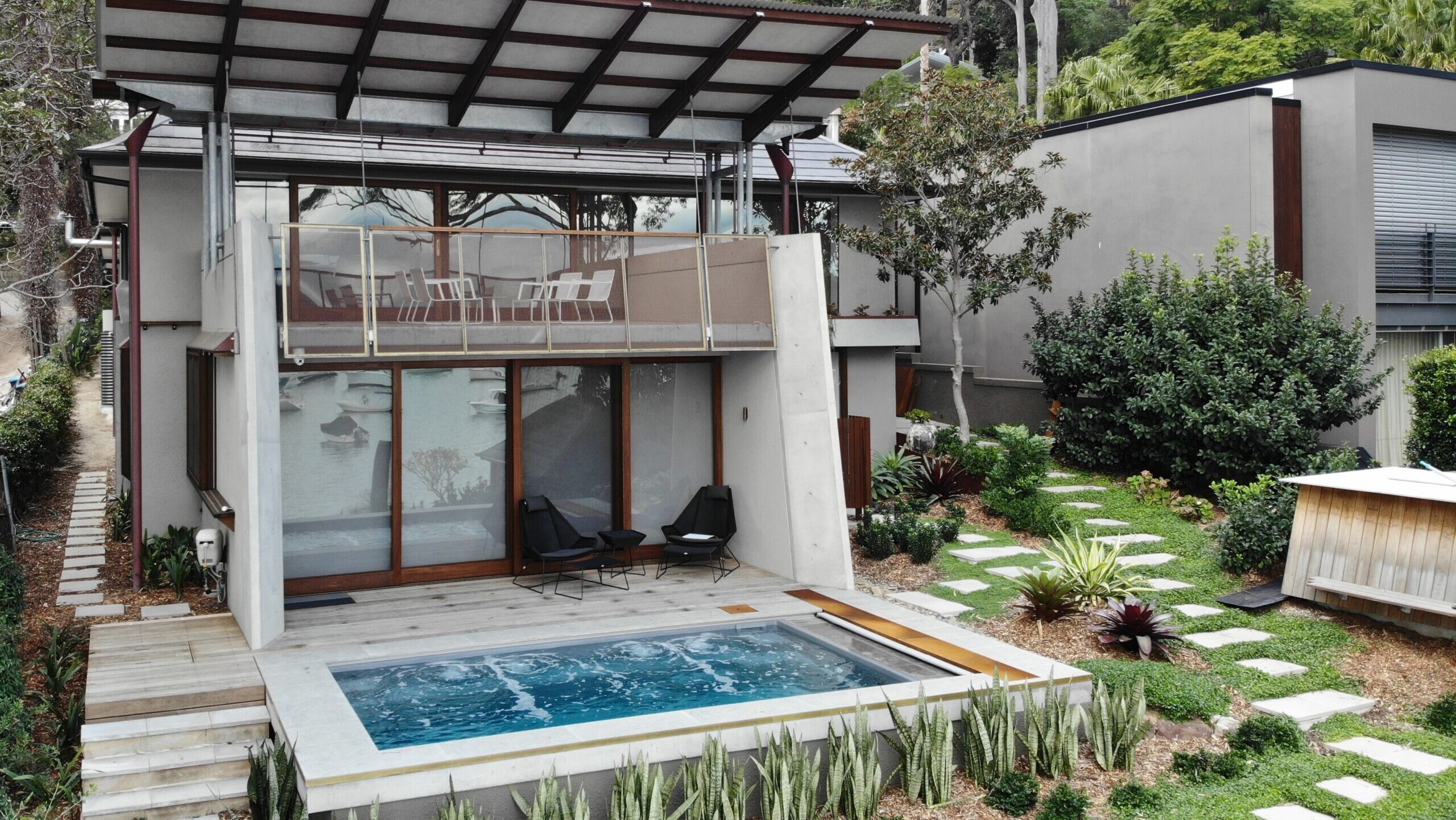 The Harmony 5.00m fibreglass swimming pool is installed in the wooden front yard of a beach house, with greenery and extra pool features surrounding it. The beautiful Graphite Grey colour stands out from the white-themed house, accented by extra yellow and grey detailing.