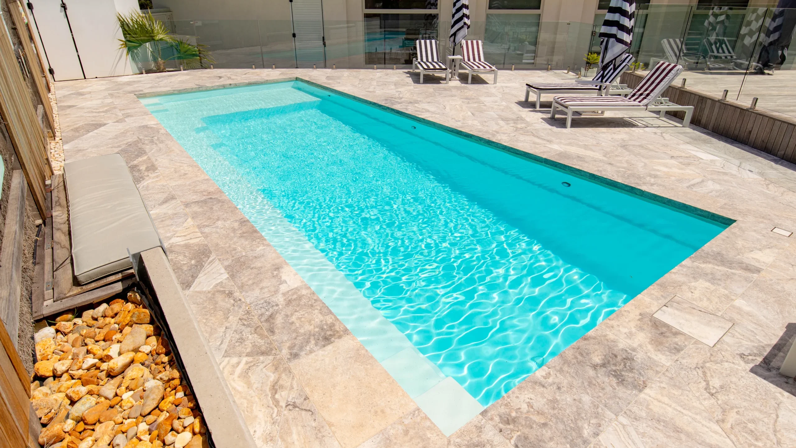 The Reflection with Splash Deck 9.50m fibreglass swimming pool is installed in the concrete backyard of a modern house, with brown tiles and greenery features surrounding it. The beautiful Shimmer White colour stands out from the white-themed house.