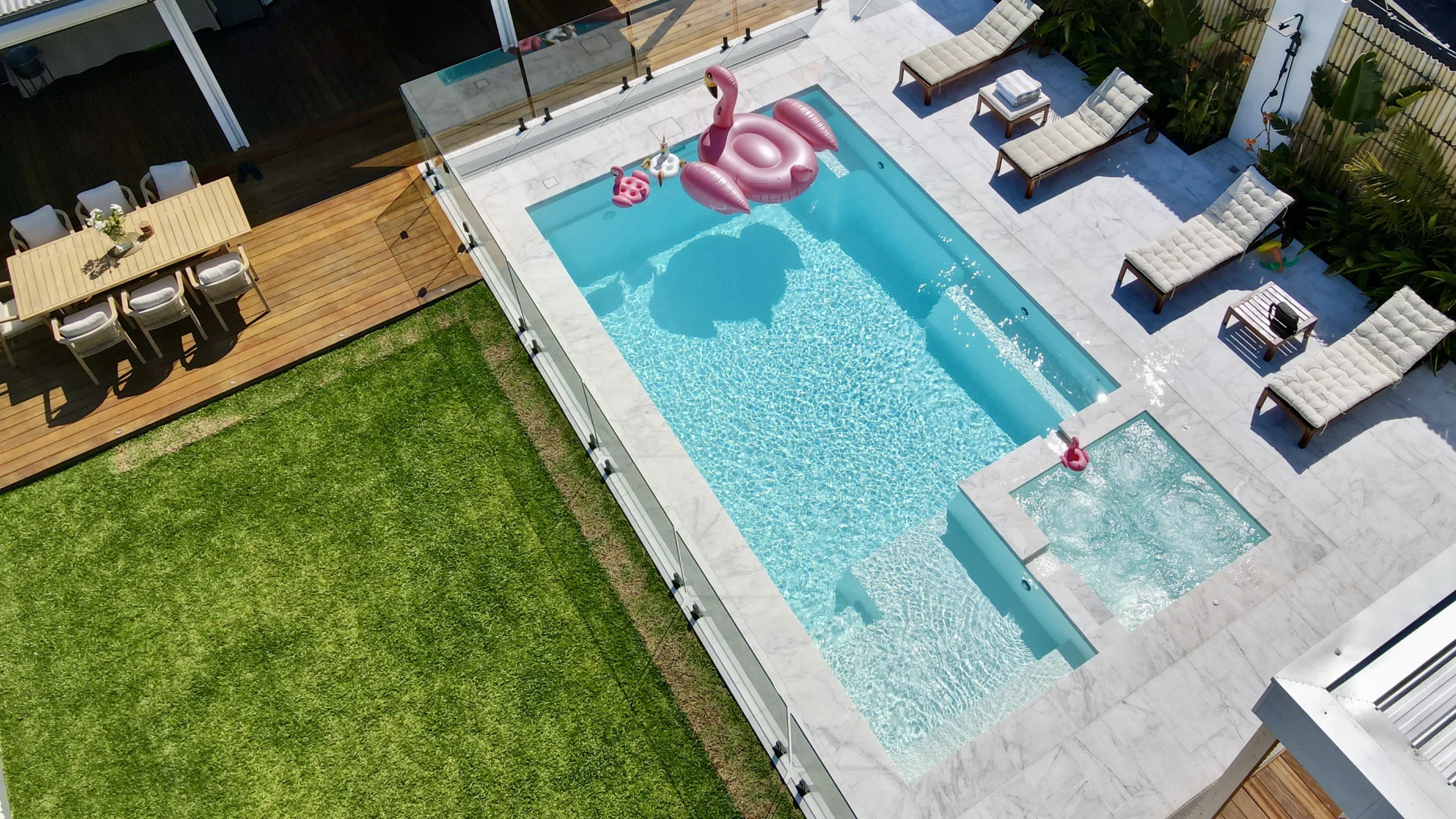 The Absolute 8.00m fibreglass swimming pool is installed in the white concrete backyard of a modern house, with greenery and extra pool features surrounding it. The beautiful Shimmer White colour stands out from the white-themed house, accented by a pink flamingo float