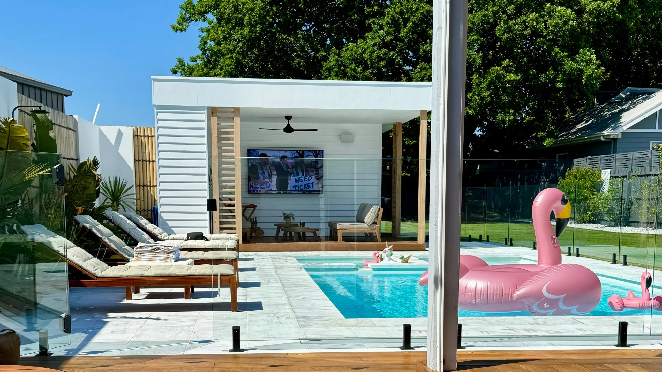 The Absolute 8.00m fibreglass swimming pool is installed in the white concrete backyard of a modern house, with greenery and extra pool features surrounding it. The beautiful Shimmer White colour stands out from the white-themed house, accented by a pink flamingo float