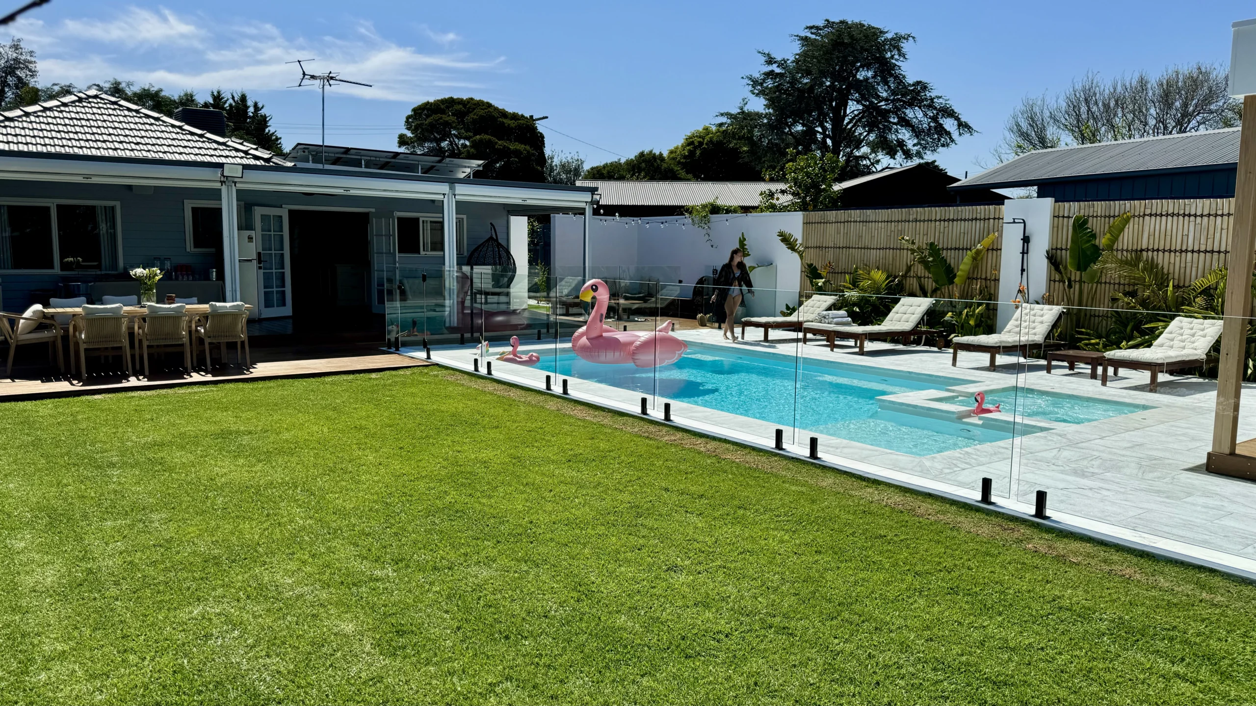 The Absolute 8.00m fibreglass swimming pool is installed in the white concrete backyard of a modern house, with greenery and extra pool features surrounding it. The beautiful Shimmer White colour stands out from the white-themed house, accented by a pink flamingo float