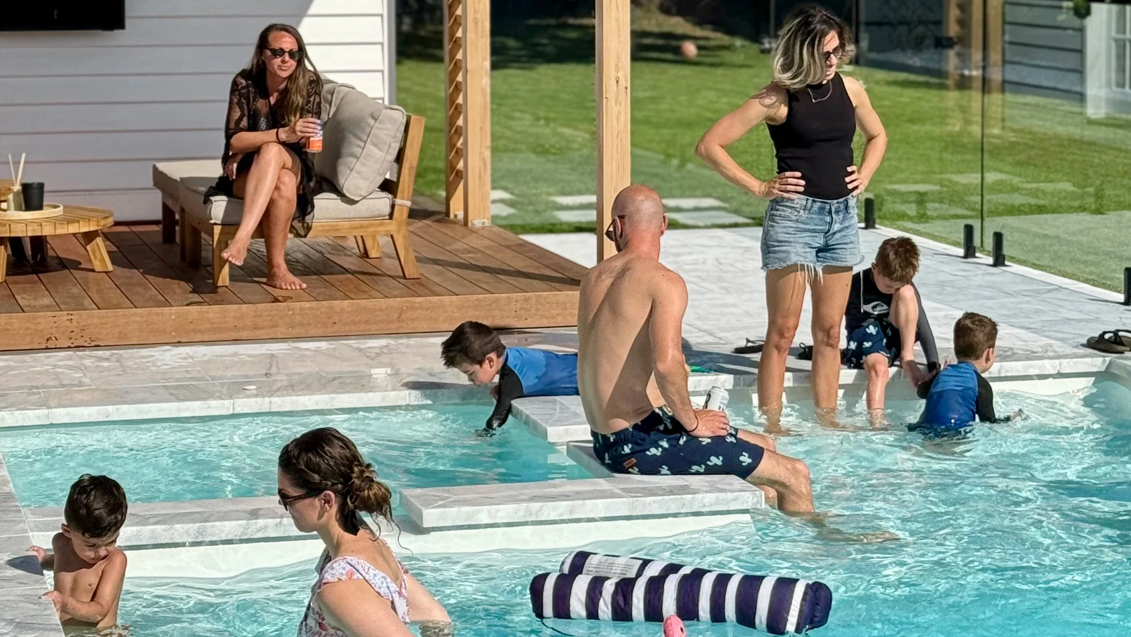 The Absolute 8.00m fibreglass swimming pool is installed in the white concrete backyard of a modern house, with greenery and extra pool features surrounding it. The beautiful Shimmer White colour stands out from the white-themed house, accented by a pink flamingo float
