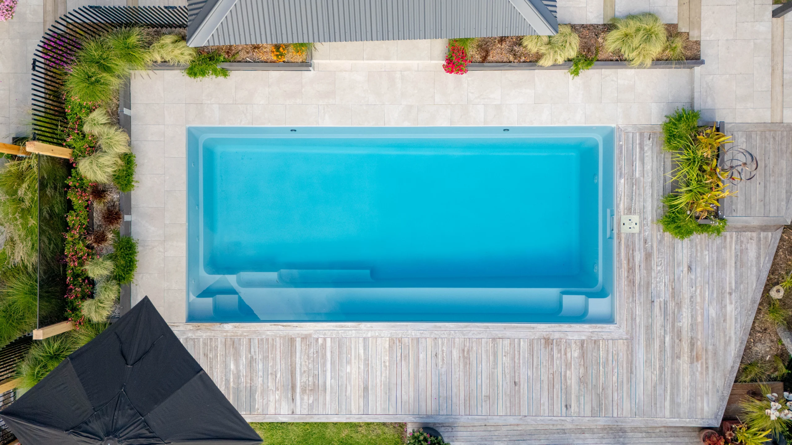 The Reflection 8.00m fibreglass swimming pool is installed in the grey concrete backyard of a modern house, with sophisticated greenery features surrounding it. The beautiful Silvery Grey colour stands out from the yellow-themed house.