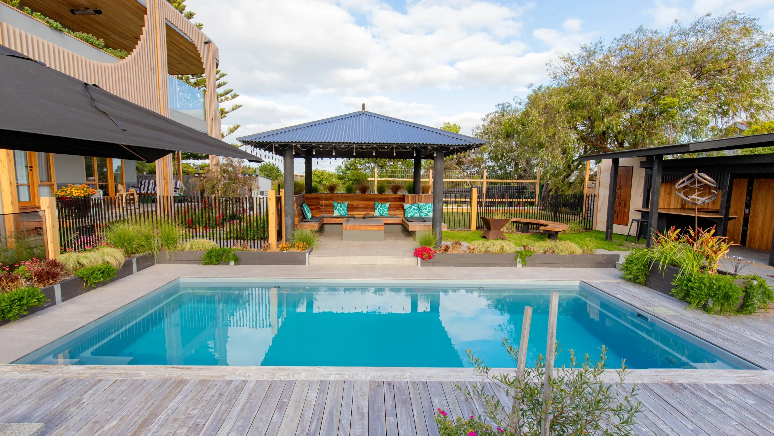 The Reflection 8.00m fibreglass swimming pool is installed in the grey concrete backyard of a modern house, with sophisticated greenery features surrounding it. The beautiful Silvery Grey colour stands out from the yellow-themed house.
