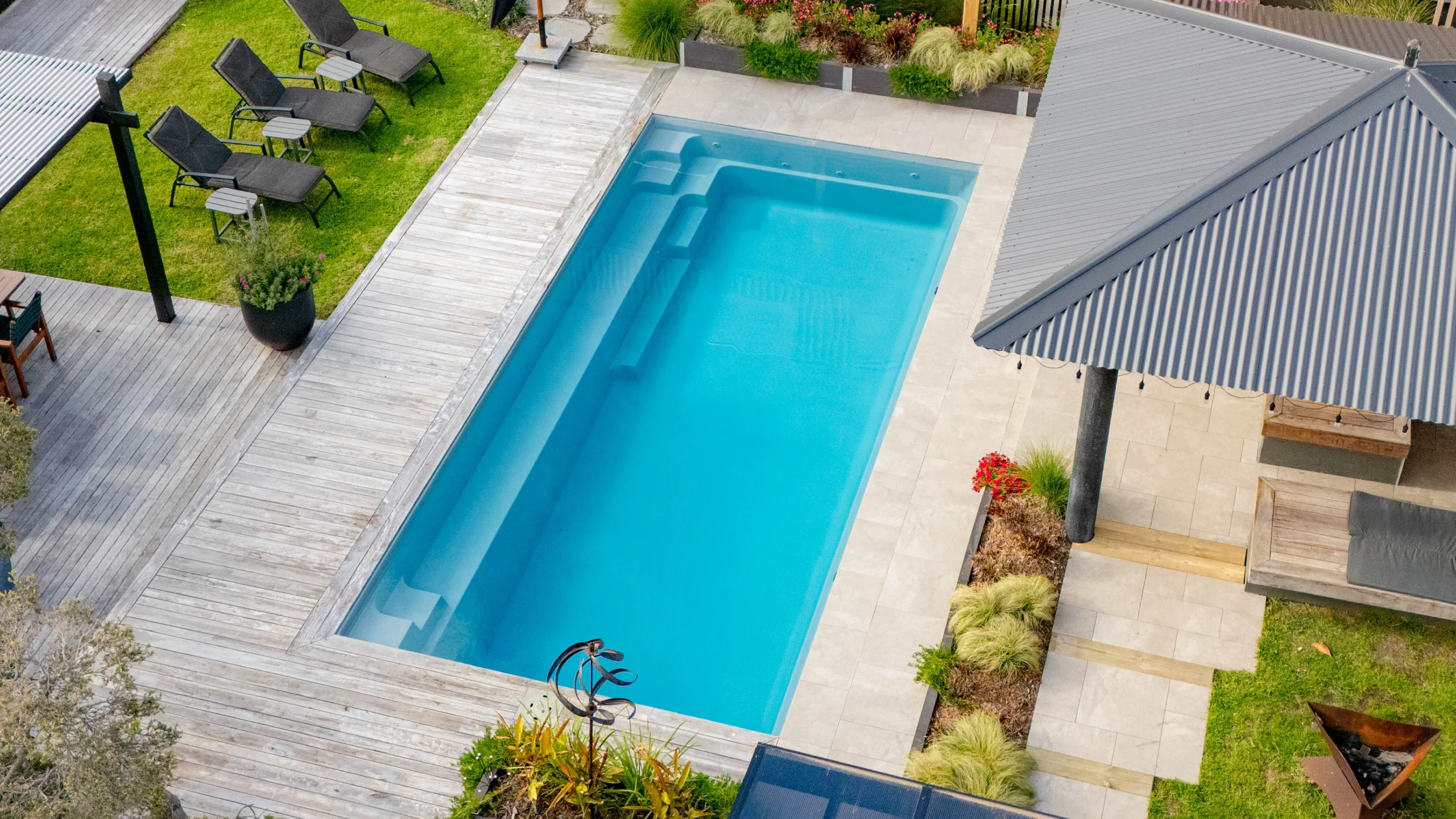 The Reflection 8.00m fibreglass swimming pool is installed in the grey concrete backyard of a modern house, with sophisticated greenery features surrounding it. The beautiful Silvery Grey colour stands out from the yellow-themed house.