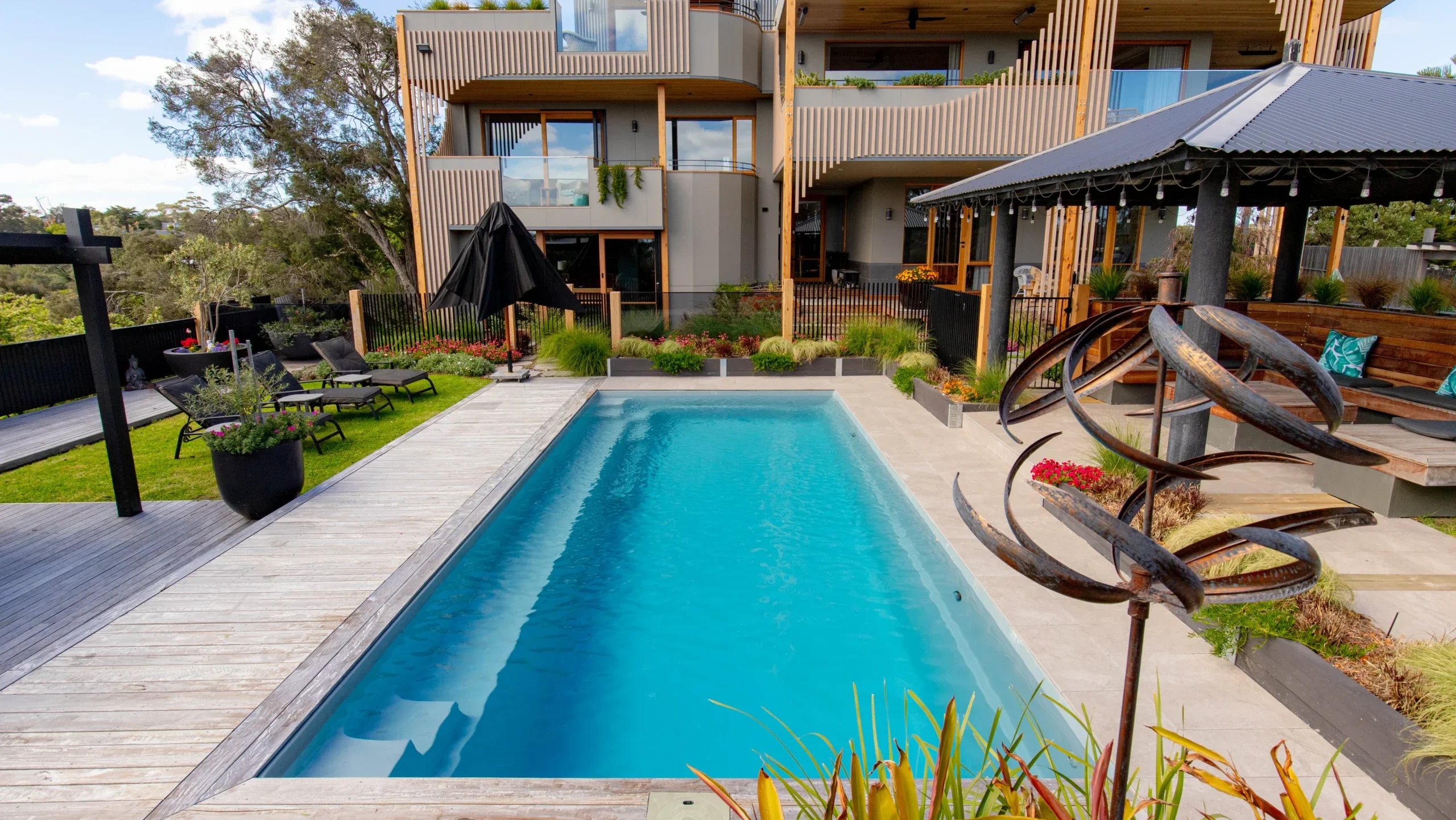 The Reflection 8.00m fibreglass swimming pool is installed in the grey concrete backyard of a modern house, with sophisticated greenery features surrounding it. The beautiful Silvery Grey colour stands out from the yellow-themed house.