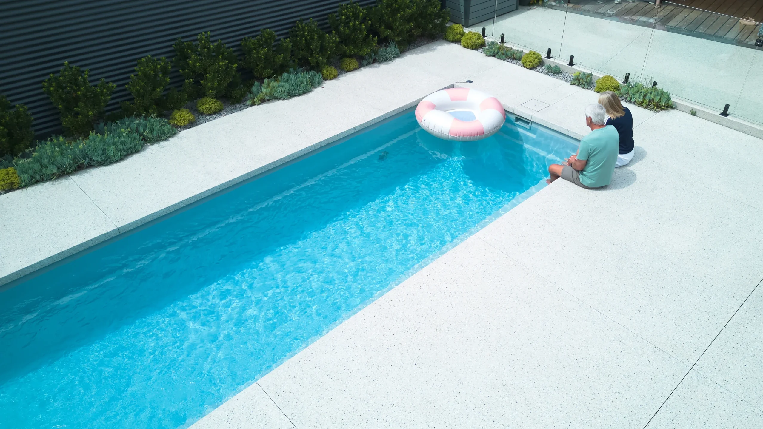 The Harmony 8.00m fibreglass swimming pool is installed in the white concrete backyard of a modern house, with greenery and extra pool features surrounding it. The beautiful silver-grey colour stands out from the white-themed house, accented by pink and blue floats.