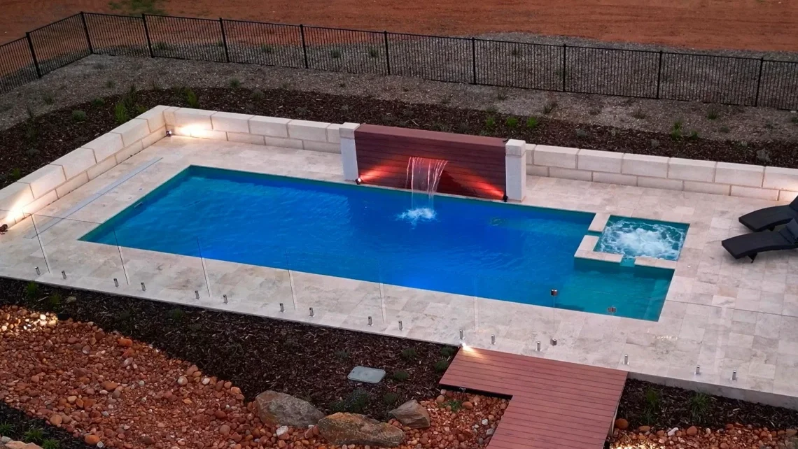 The Absolute 12.00m fibreglass swimming pool with a built-in spa is in the middle of the backyard, with red tiles and greenery surrounding it. The pool colour is aquamarine makes the fibreglass swimming pool stand out from the mountainous background and dawn sky.