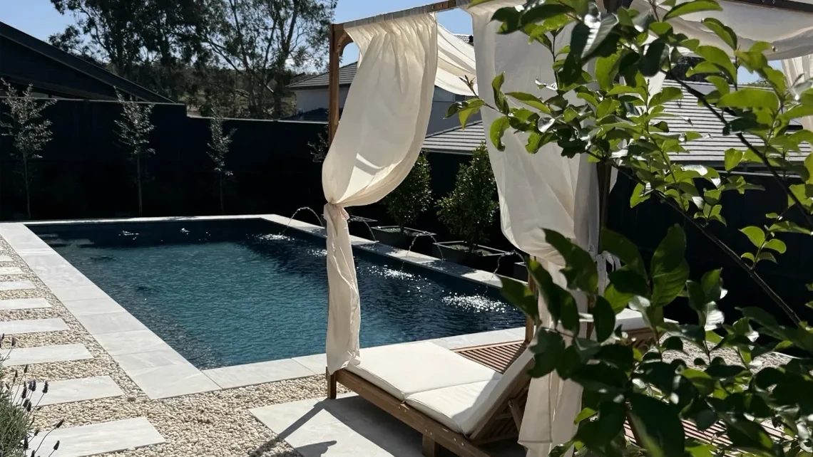 The Reflection 8.00m fibreglass swimming pool is installed in the white concrete backyard of a modern house, with lavenders and extra pool features surrounding it. The beautiful Graphite Grey colour stands out from the white-themed house, accented by a pair of white pool curtains.