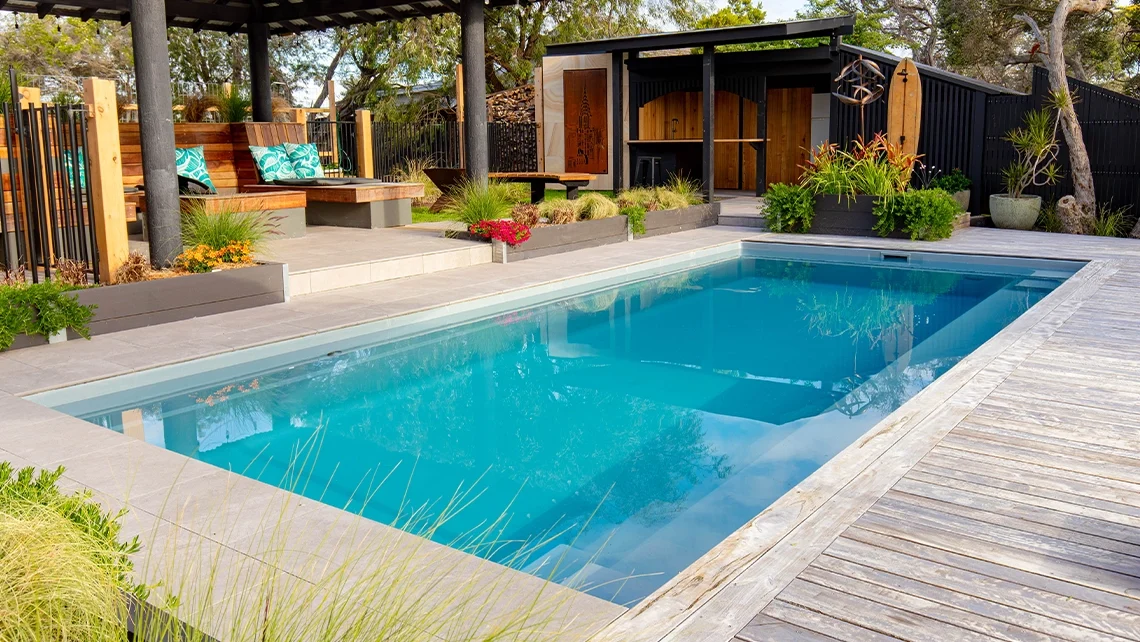 The Reflection 8.00m fibreglass swimming pool is installed in the grey concrete backyard of a modern house, with sophisticated greenery features surrounding it. The beautiful Silvery Grey colour stands out from the yellow-themed house.