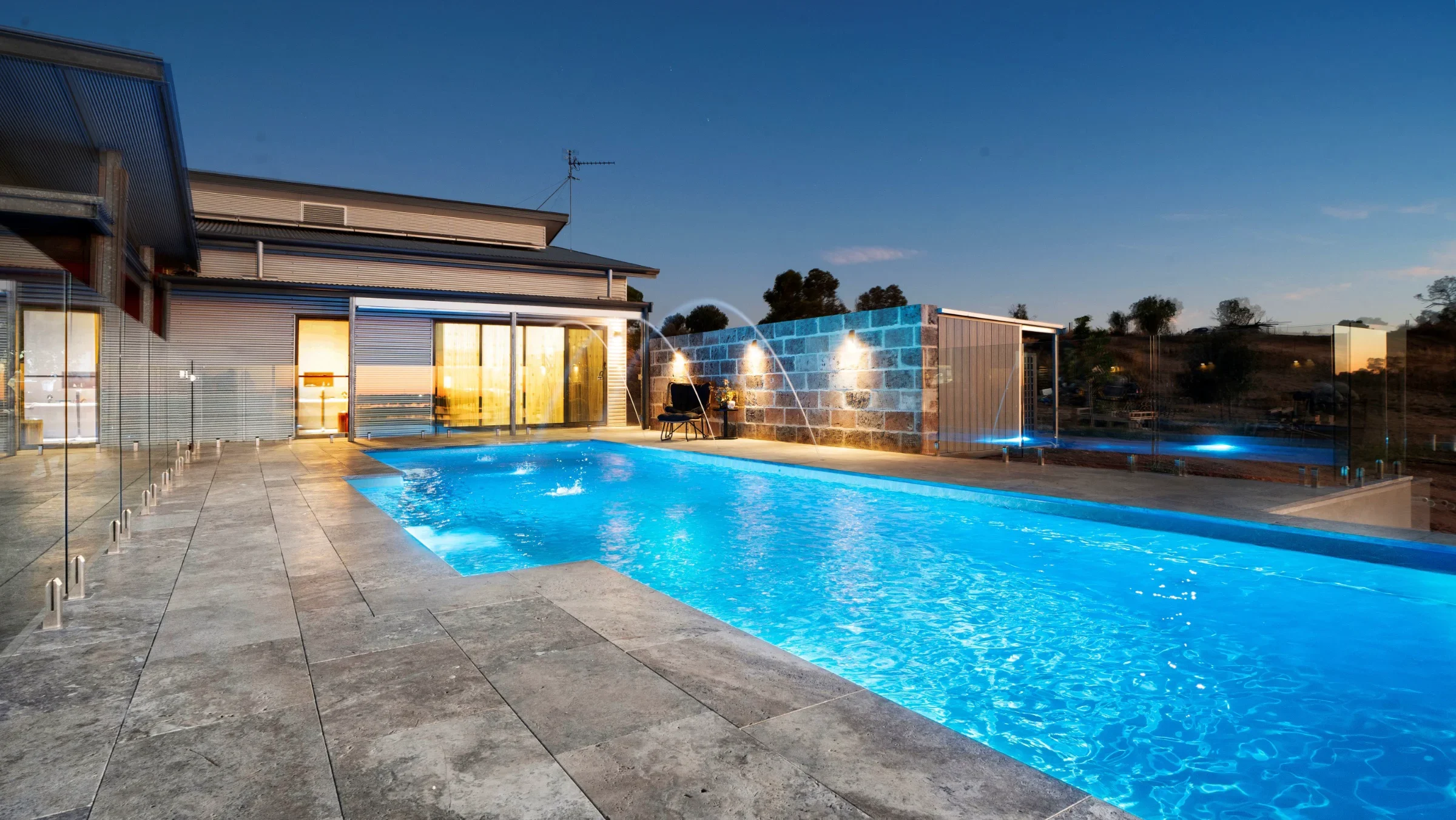 The Elegance 12.00m fibreglass swimming pool is installed in an ocean-viewing backyard of a modern house, with grey tiles and waterfall features surrounding it. The beautiful Crystal Blue colour shines brightly against the orange dawn sky in the backyard.