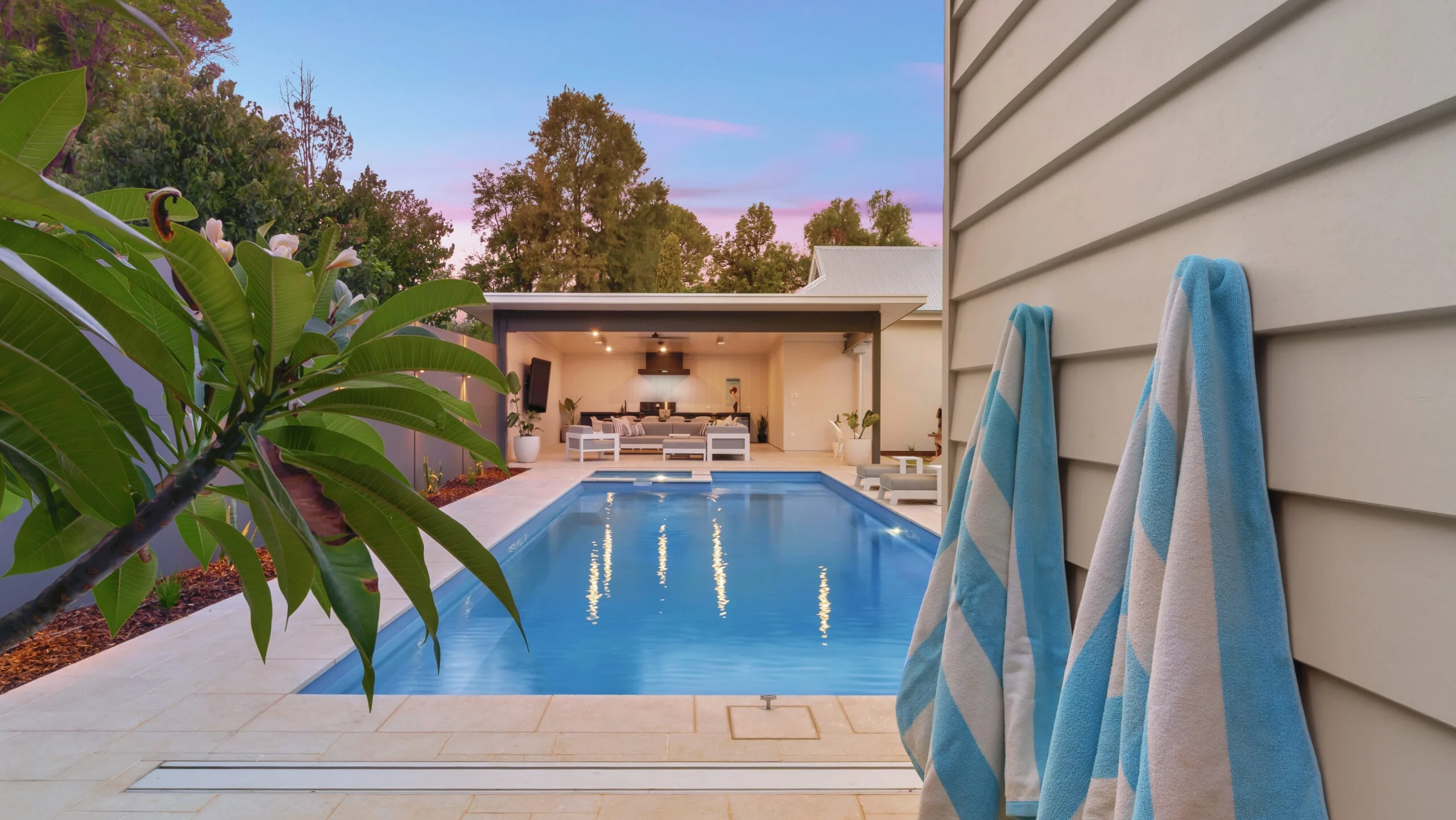 The Absolute 12.00m fibreglass swimming pool is installed in the heart of a heritage house with a pool house next to it and palm trees in the background. Its stunning Silver Grey colour stands out from the white heritage house with dim lighting at dawn.