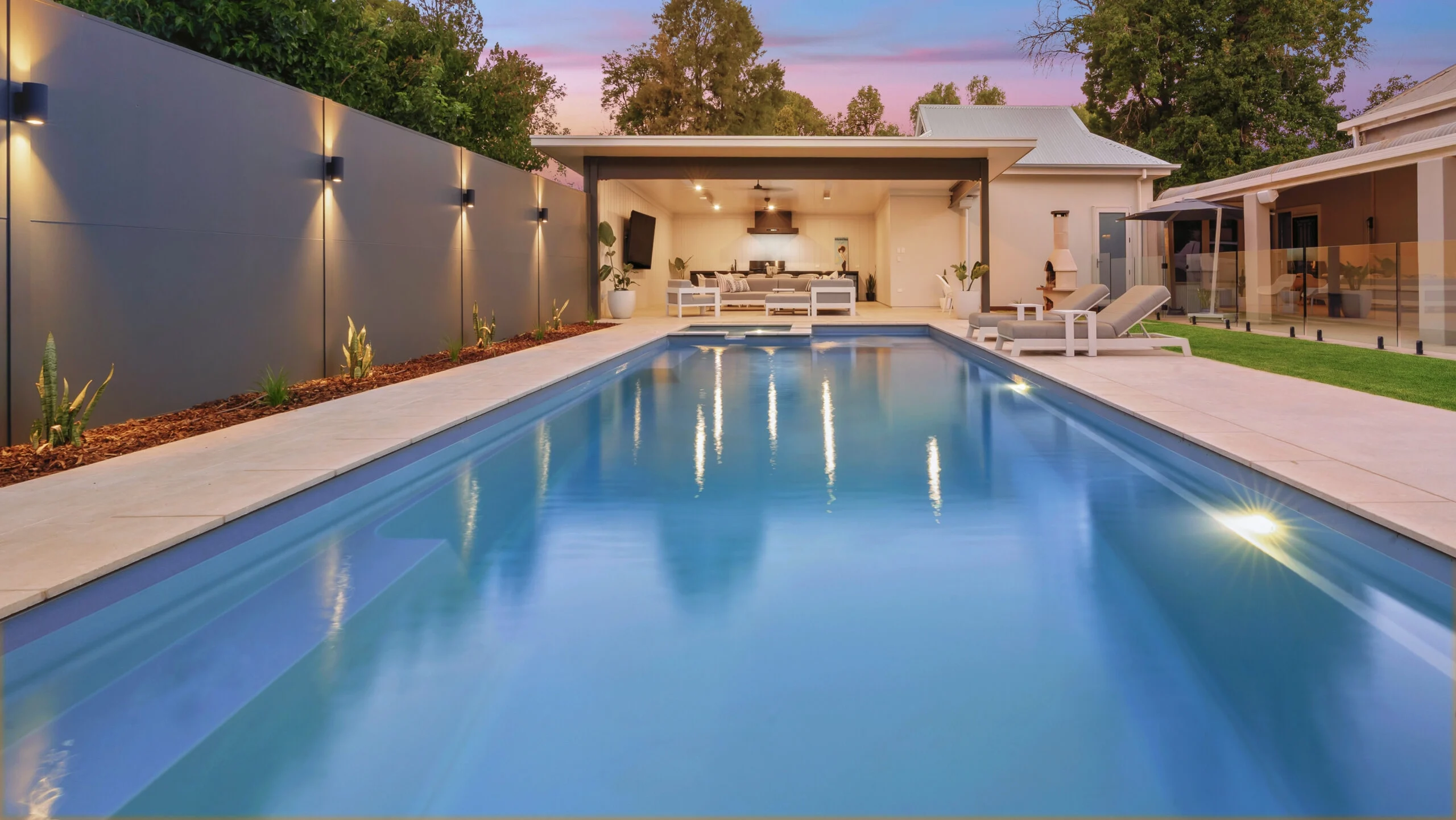 The Absolute 12.00m fibreglass swimming pool is installed in the heart of a heritage house with a pool house next to it and palm trees in the background. Its stunning Silver Grey colour stands out from the white heritage house with dim lighting at dawn.