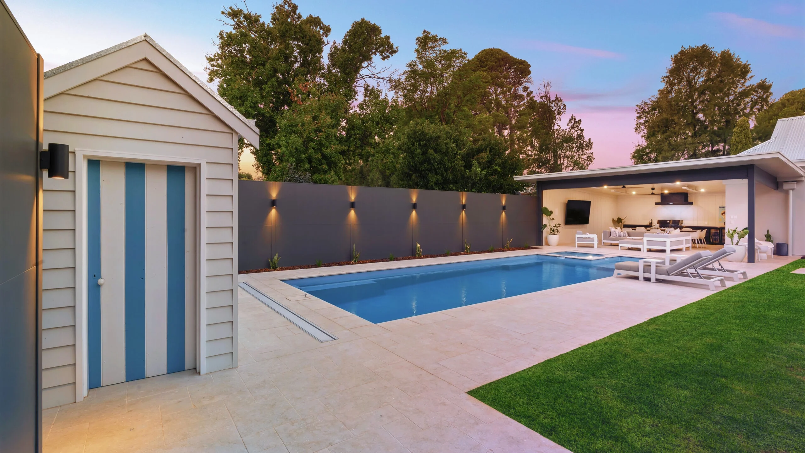 The Absolute 12.00m fibreglass swimming pool is installed in the heart of a heritage house with a pool house next to it and palm trees in the background. Its stunning Silver Grey colour stands out from the white heritage house with dim lighting at dawn.