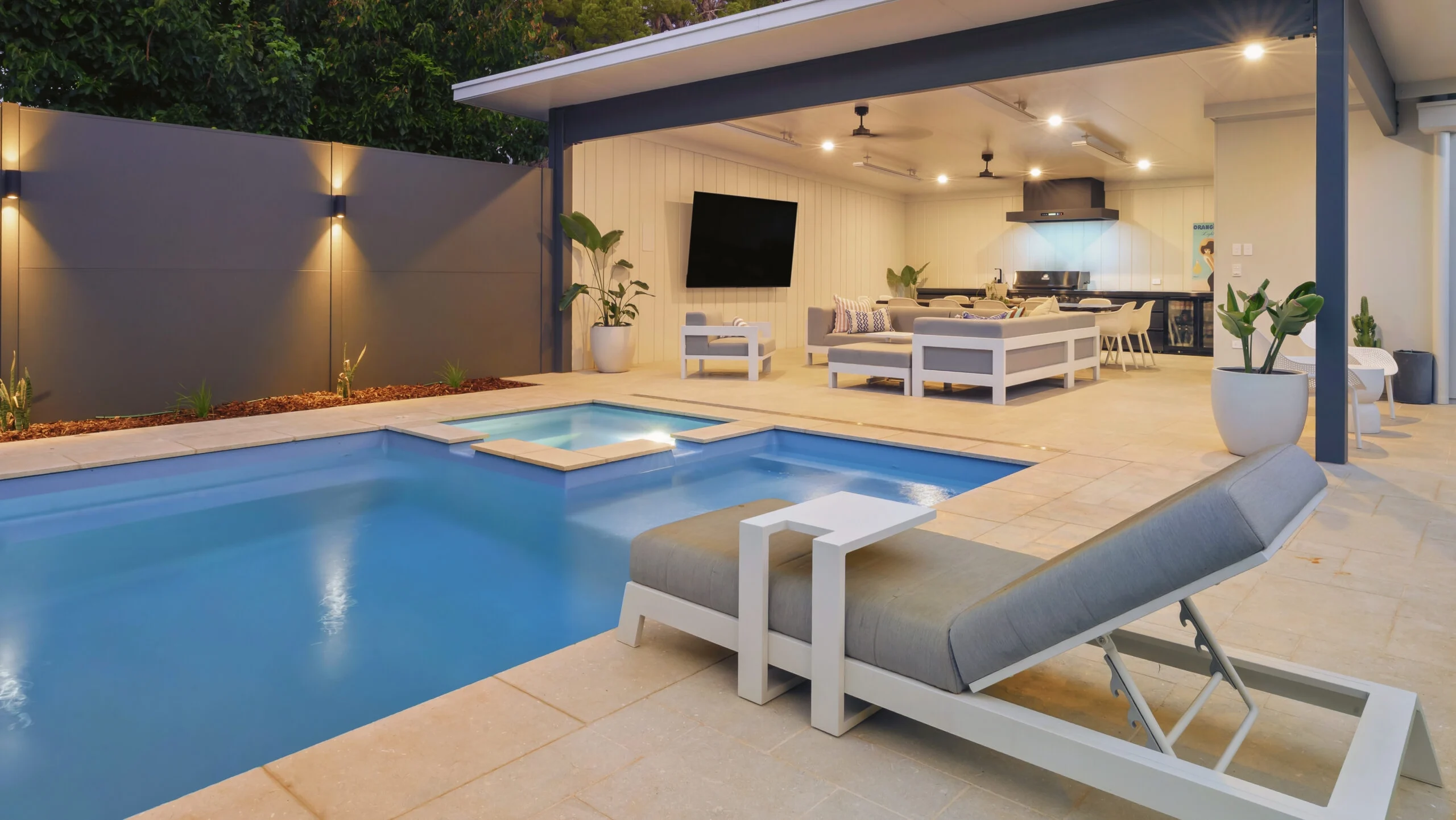 The Absolute 12.00m fibreglass swimming pool is installed in the heart of a heritage house with a pool house next to it and palm trees in the background. Its stunning Silver Grey colour stands out from the white heritage house with dim lighting at dawn.