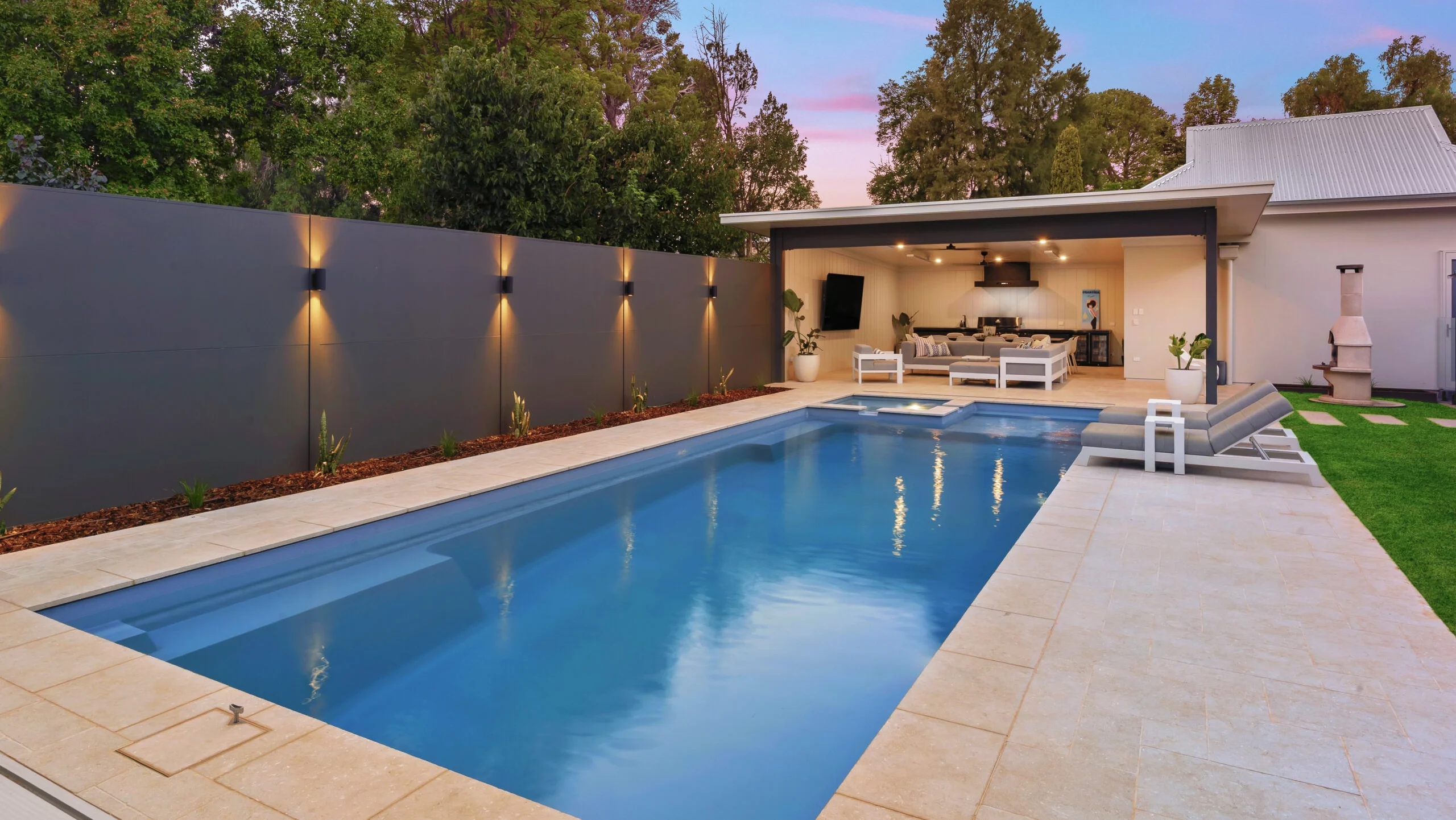 The Absolute 12.00m fibreglass swimming pool is installed in the heart of a heritage house with a pool house next to it and palm trees in the background. Its stunning Silver Grey colour stands out from the white heritage house with dim lighting at dawn.