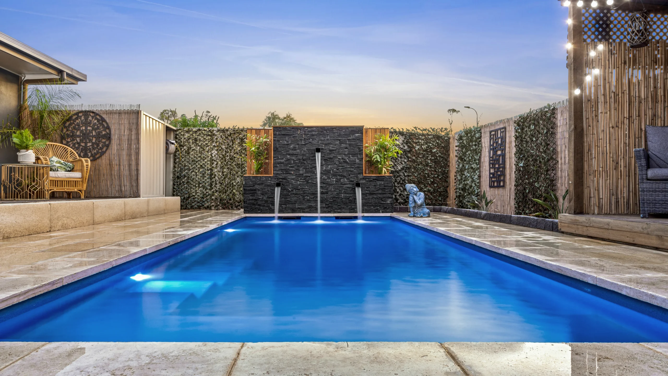 The Precision 8.00m fibreglass swimming pool is installed in a serene backyard of a modern house, with grey tiles and waterfall features surrounding it. The beautiful Sapphire Blue colour merges seamlessly with the blue sky in the backyard.