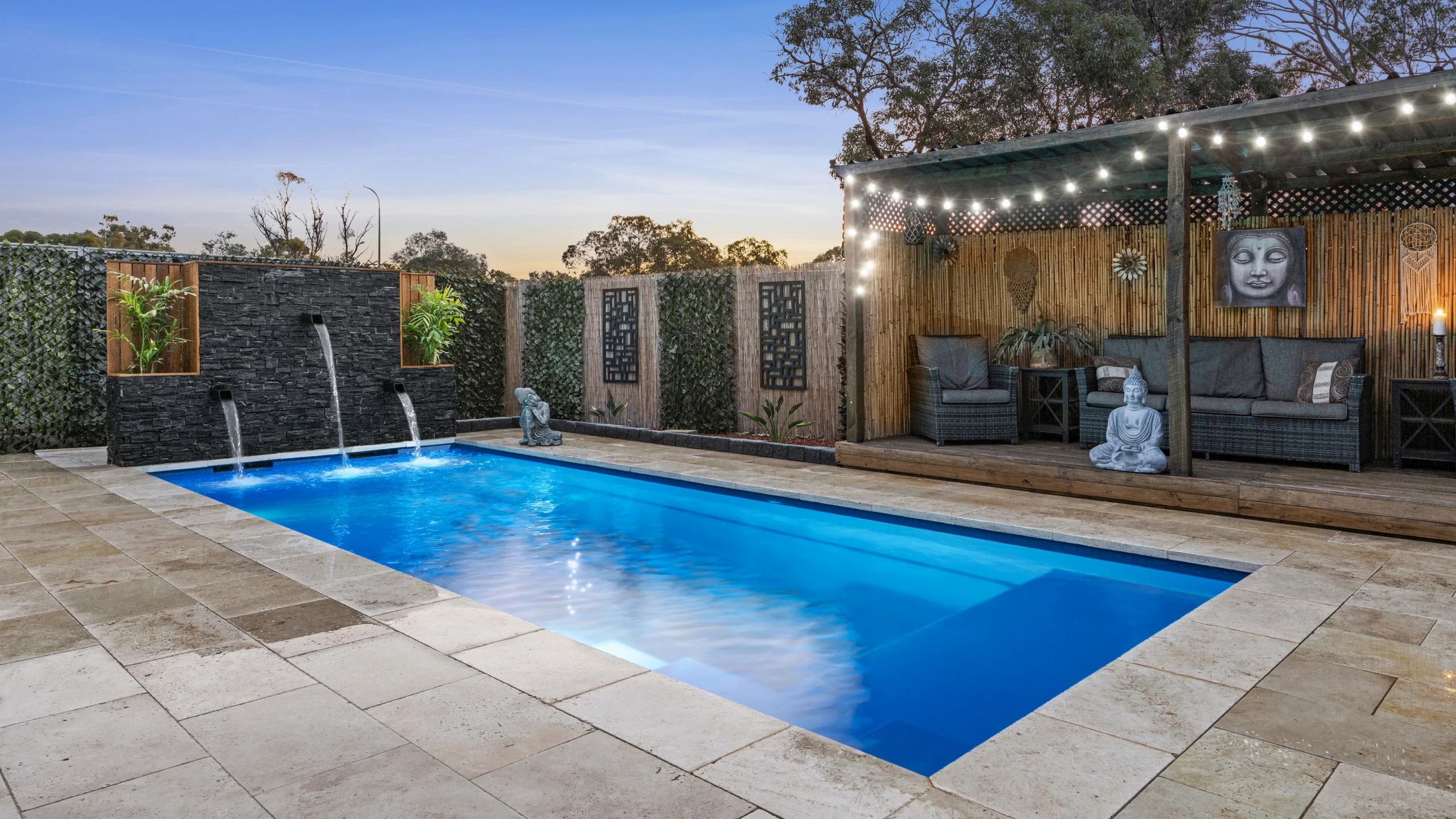 The Precision 8.00m fibreglass swimming pool is installed in a serene backyard of a modern house, with grey tiles and waterfall features surrounding it. The beautiful Sapphire Blue colour merges seamlessly with the blue sky in the backyard.