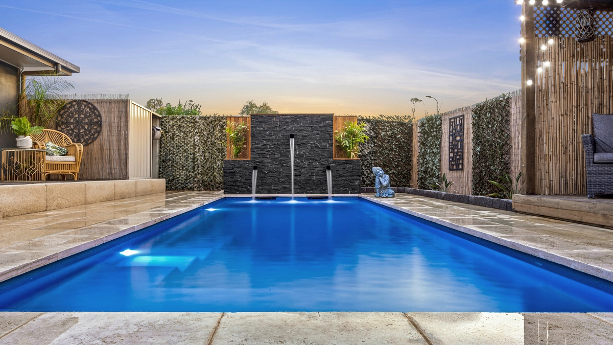 The Precision 8.00m fibreglass swimming pool is installed in a serene backyard of a modern house, with grey tiles and waterfall features surrounding it. The beautiful Sapphire Blue colour merges seamlessly with the blue sky in the backyard.