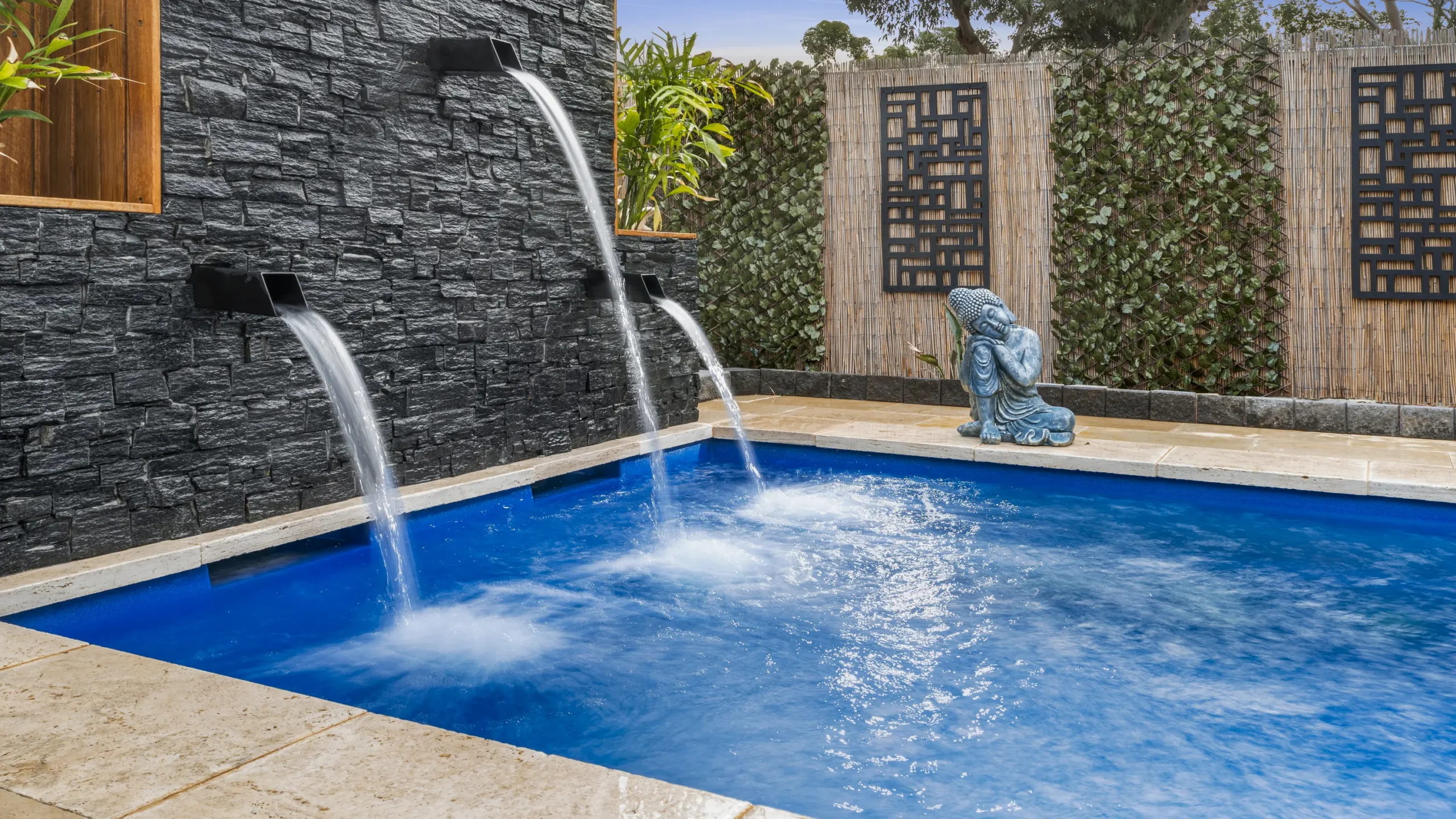The Precision 8.00m fibreglass swimming pool is installed in a serene backyard of a modern house, with grey tiles and waterfall features surrounding it. The beautiful Sapphire Blue colour merges seamlessly with the blue sky in the backyard.