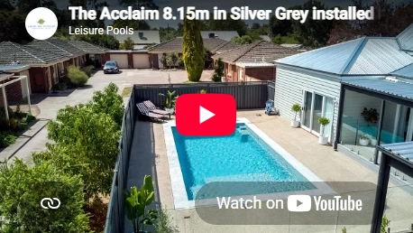 The Acclaim 8.15m fibreglass swimming pool is installed in a concrete backyard of a modern house, with grey tiles and greenery features surrounding it. The beautiful Silver Grey colour stands out from the blue house.