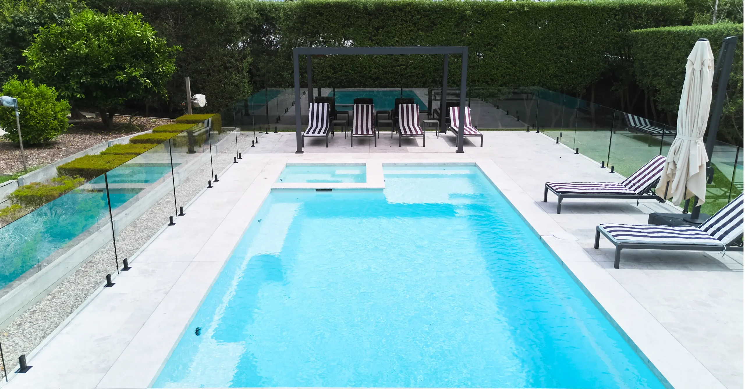The Absolute 8.00m fibreglass swimming pool in Shimmer White is installed in a landscaped backyard in Rye, Mornington Peninsula, Victoria. The pool is surrounded by light stone paving, frameless glass fencing, manicured hedges and a shaded outdoor seating area. The Shimmer White finish creates bright, pale blue water that complements the neutral paving and lush greenery, creating a fresh and inviting space for family swimming and relaxation.