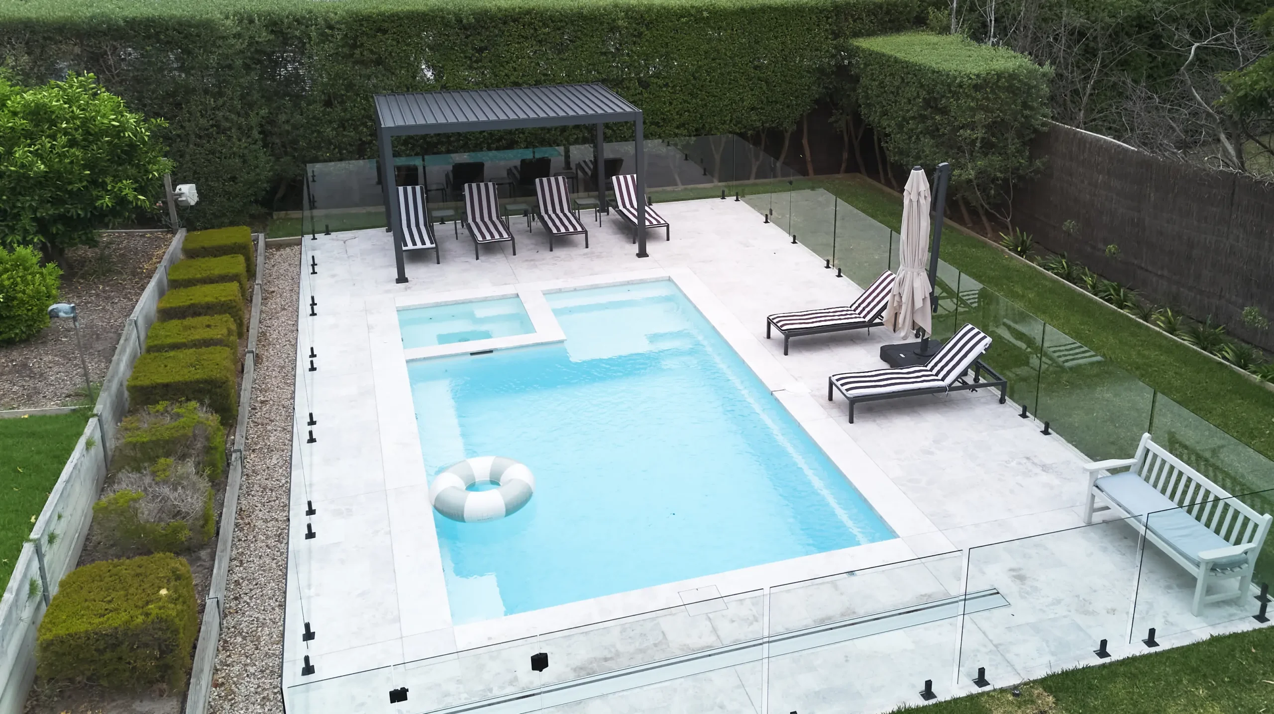 The Absolute 8.00m fibreglass swimming pool in Shimmer White is installed in a landscaped backyard in Rye, Mornington Peninsula, Victoria. The pool is surrounded by light stone paving, frameless glass fencing, manicured hedges and a shaded outdoor seating area. The Shimmer White finish creates bright, pale blue water that complements the neutral paving and lush greenery, creating a fresh and inviting space for family swimming and relaxation.