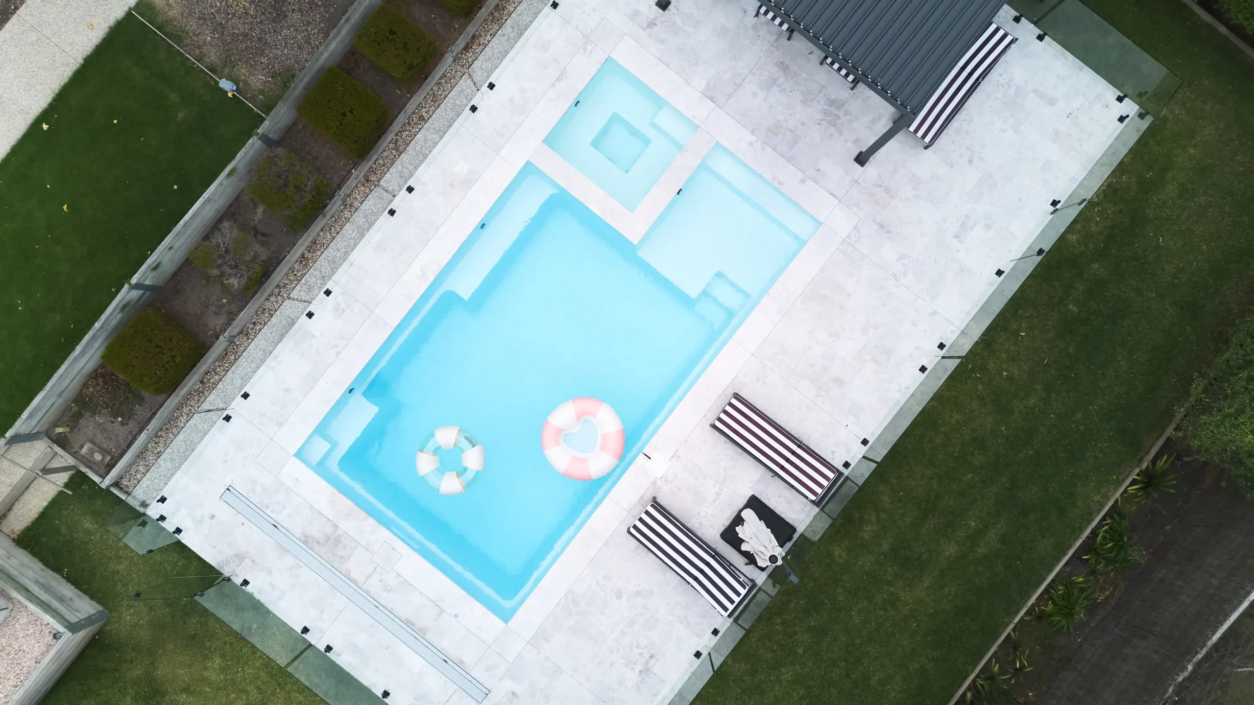 The Absolute 8.00m fibreglass swimming pool in Shimmer White is installed in a landscaped backyard in Rye, Mornington Peninsula, Victoria. The pool is surrounded by light stone paving, frameless glass fencing, manicured hedges and a shaded outdoor seating area. The Shimmer White finish creates bright, pale blue water that complements the neutral paving and lush greenery, creating a fresh and inviting space for family swimming and relaxation.