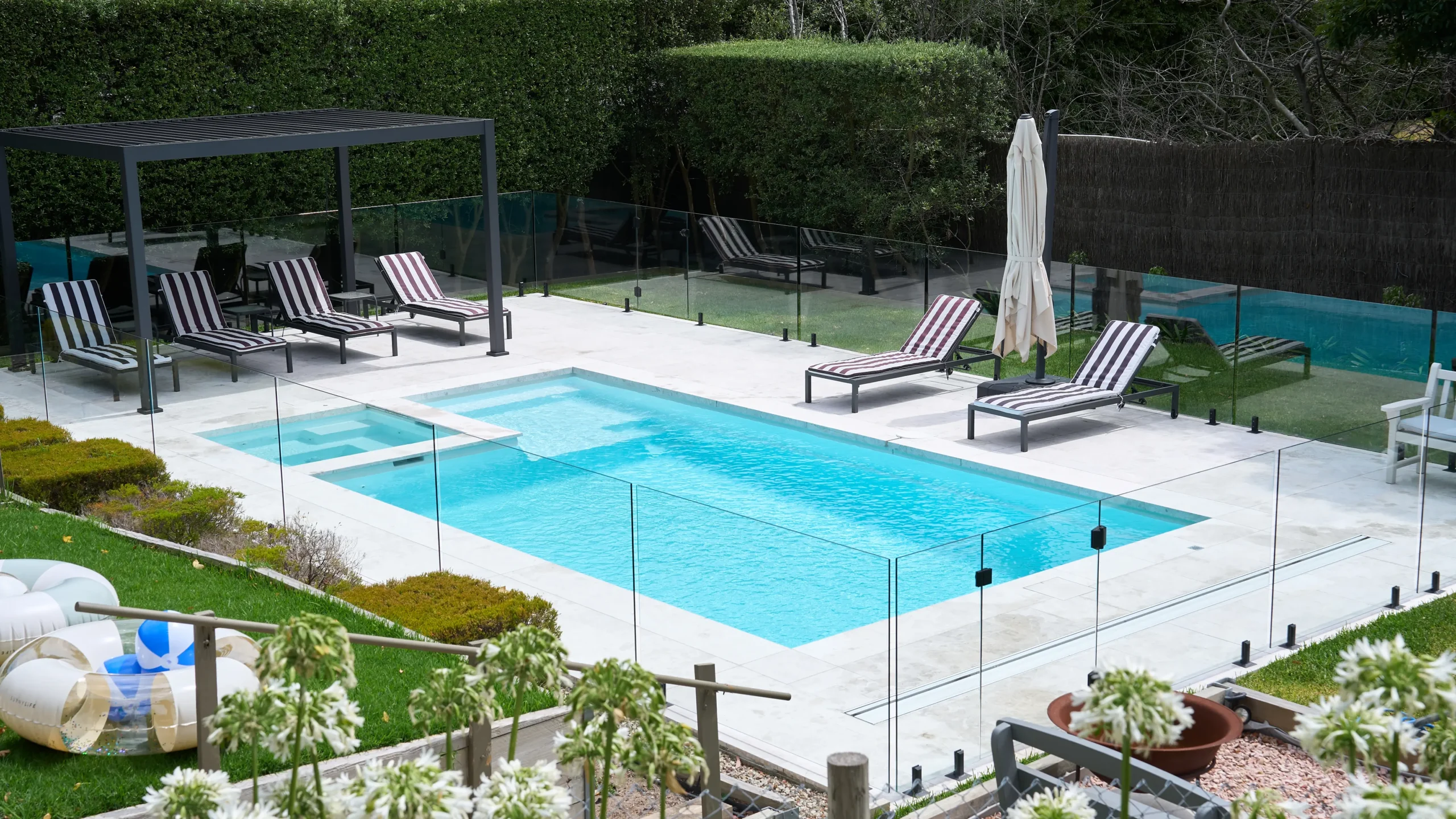 The Absolute 8.00m fibreglass swimming pool in Shimmer White is installed in a landscaped backyard in Rye, Mornington Peninsula, Victoria. The pool is surrounded by light stone paving, frameless glass fencing, manicured hedges and a shaded outdoor seating area. The Shimmer White finish creates bright, pale blue water that complements the neutral paving and lush greenery, creating a fresh and inviting space for family swimming and relaxation.