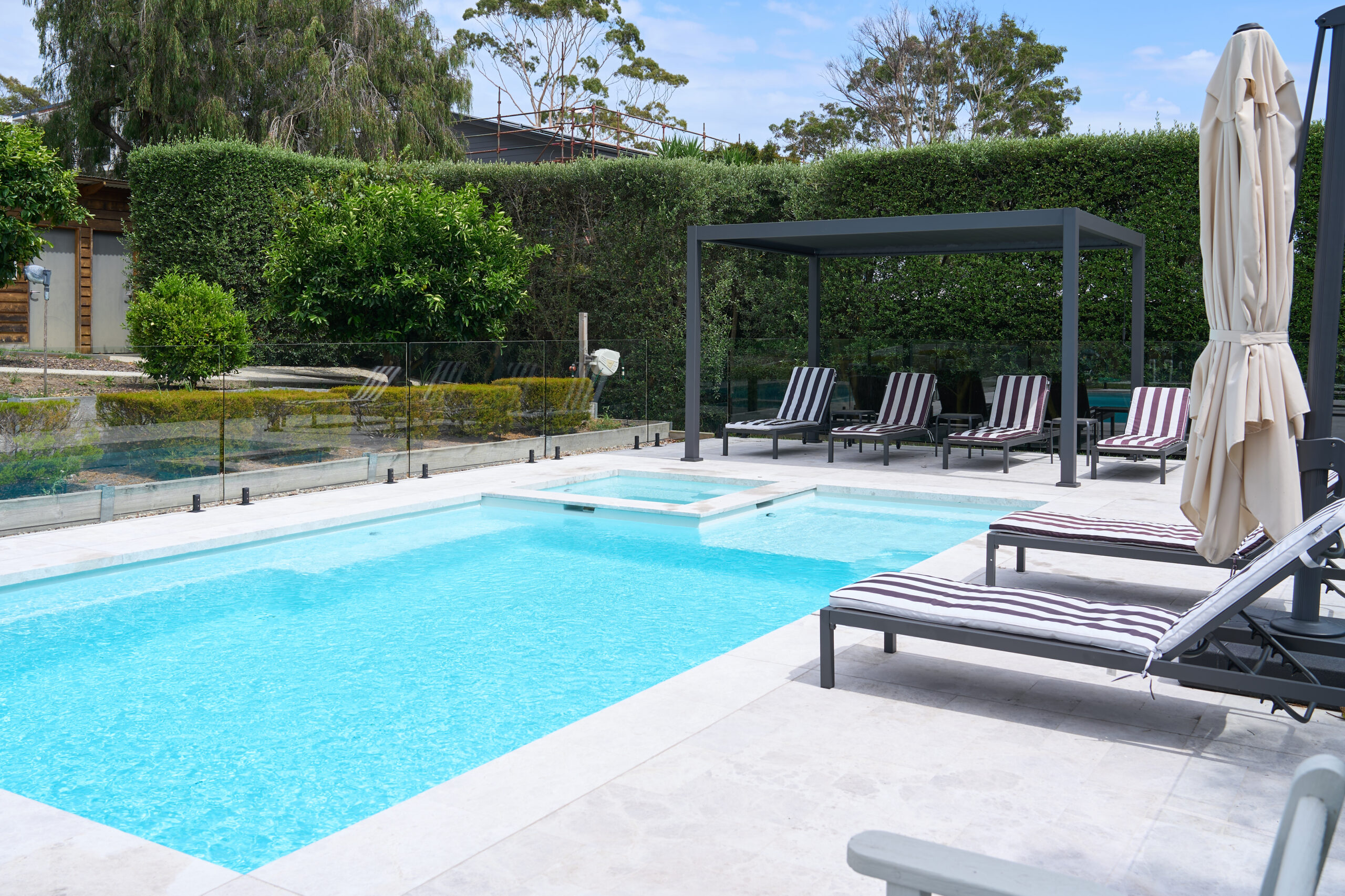 The Absolute 8.00m fibreglass swimming pool in Shimmer White is installed in a landscaped backyard in Rye, Mornington Peninsula, Victoria. The pool is surrounded by light stone paving, frameless glass fencing, manicured hedges and a shaded outdoor seating area. The Shimmer White finish creates bright, pale blue water that complements the neutral paving and lush greenery, creating a fresh and inviting space for family swimming and relaxation.