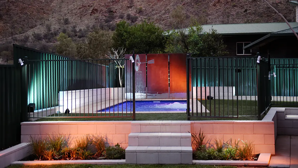The Harmony 7.00m fibreglass swimming pool is installed in the gated backyard of a mountainous house with white concrete tiles and a windmill surrounding it. The beautiful Sapphire Blue pool colour stands out from the white-themed house and grey dawn sky.