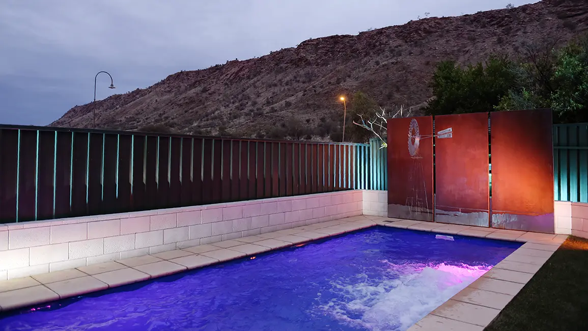 The Harmony 7.00m fibreglass swimming pool is installed in the gated backyard of a mountainous house with white concrete tiles and a windmill surrounding it. The beautiful Sapphire Blue pool colour stands out from the white-themed house and grey dawn sky.