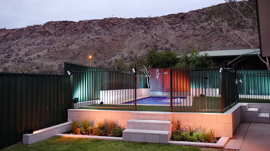 The Harmony 7.00m fibreglass swimming pool is installed in the gated backyard of a mountainous house with white concrete tiles and a windmill surrounding it. The beautiful Sapphire Blue pool colour stands out from the white-themed house and grey dawn sky.