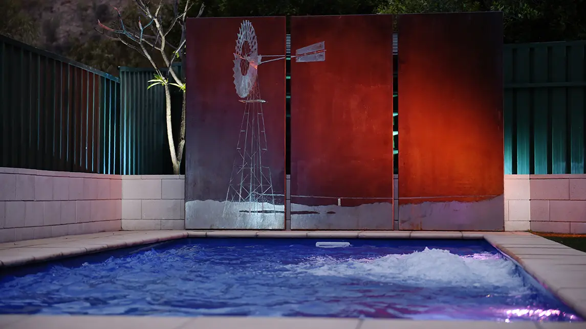 The Harmony 7.00m fibreglass swimming pool is installed in the gated backyard of a mountainous house with white concrete tiles and a windmill surrounding it. The beautiful Sapphire Blue pool colour stands out from the white-themed house and grey dawn sky.