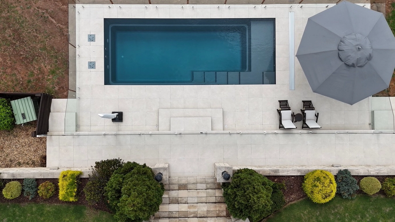 The Precision 8.05m fibreglass swimming pool is installed in front of a large white countryside mansion, with greenery and mountains surrounding it. The mysterious Graphite Grey gives a modern look to the overall countryside aesthetics.