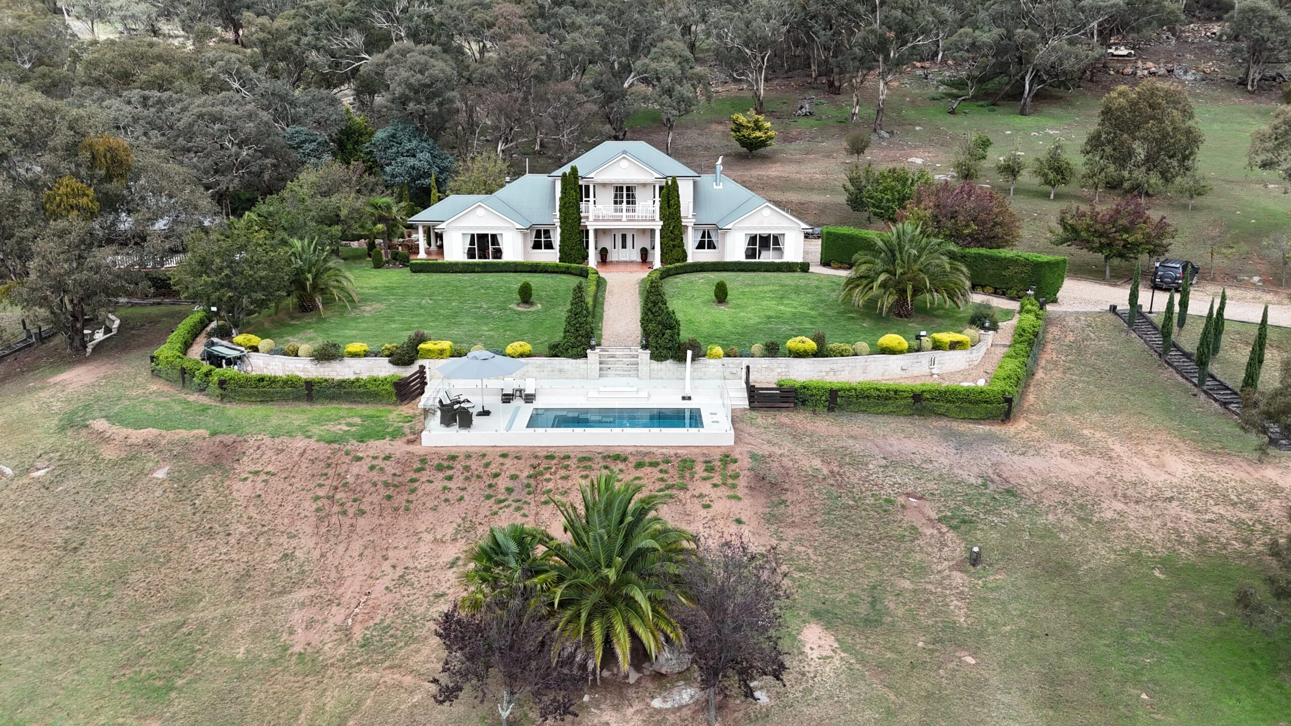 The Precision 8.05m fibreglass swimming pool is installed in front of a large white countryside mansion, with greenery and mountains surrounding it. The mysterious Graphite Grey gives a modern look to the overall countryside aesthetics.
