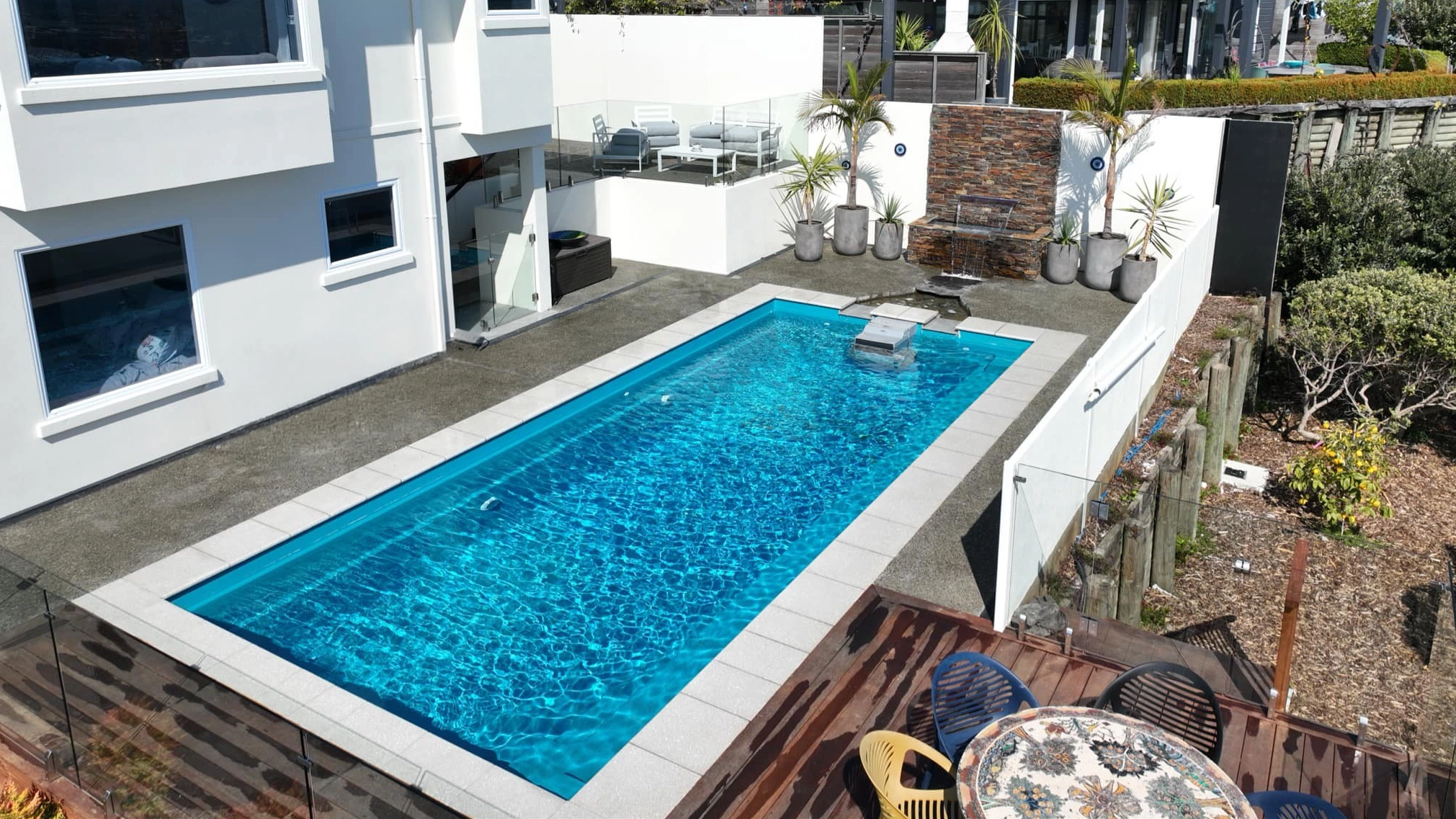 IMG LP NZ Reflection 9 w AC Crystal Blue NP Nelson 002 The Reflection with Auto Cover 9.90m fibreglass swimming pool is installed in a metropolitan region of Stoke, New Zealand. The house is surrounded by grey tiles, a small waterfall and natural greenery with mountains in the background. The pool colour is Crystal Blue, which stands out from the tranquil landscape and white house.