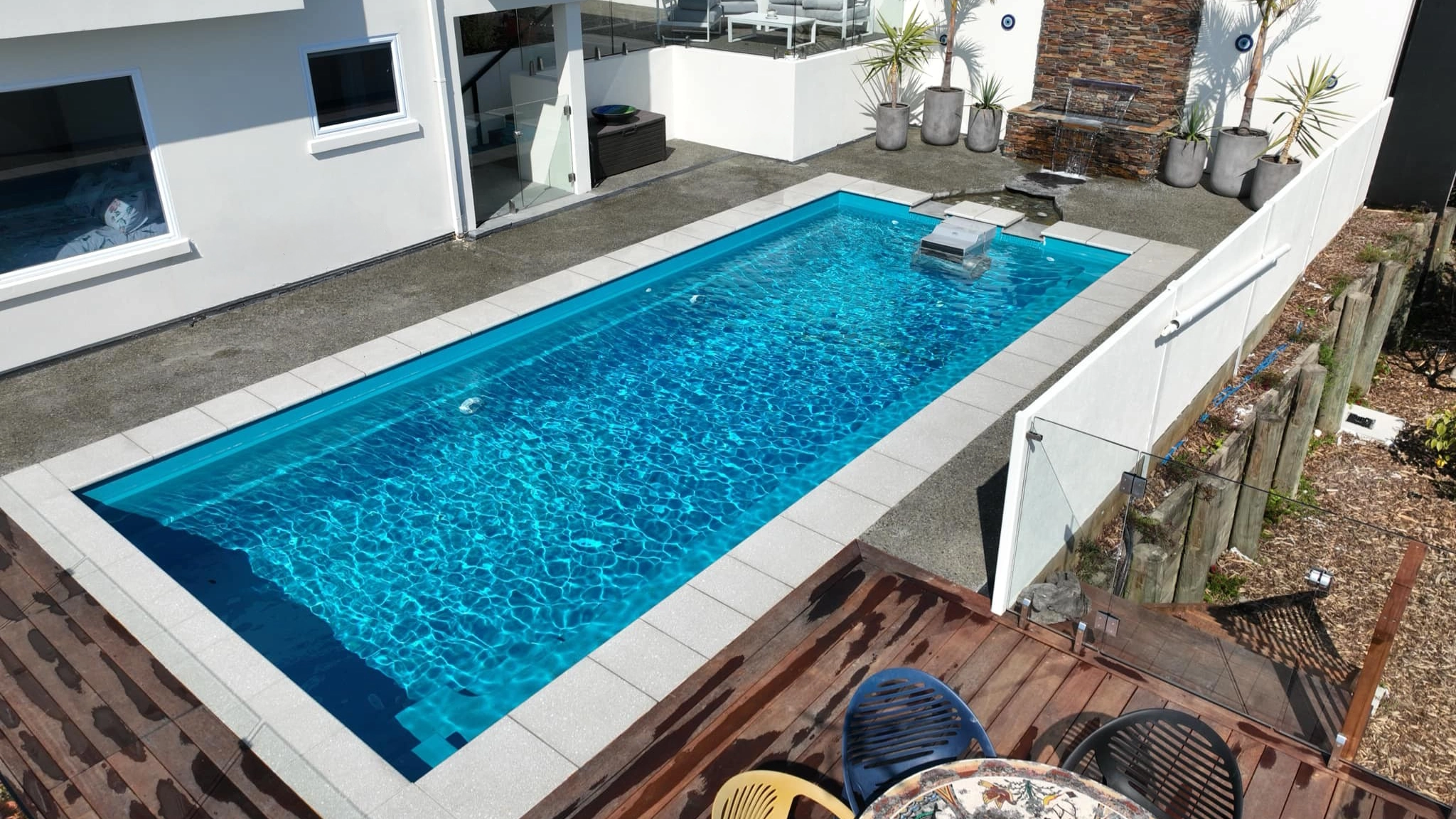 The Reflection with Auto Cover 9.90m fibreglass swimming pool is installed in a metropolitan region of Stoke, New Zealand. The house is surrounded by grey tiles, a small waterfall and natural greenery with mountains in the background. The pool colour is Crystal Blue, which stands out from the tranquil landscape and white house.
