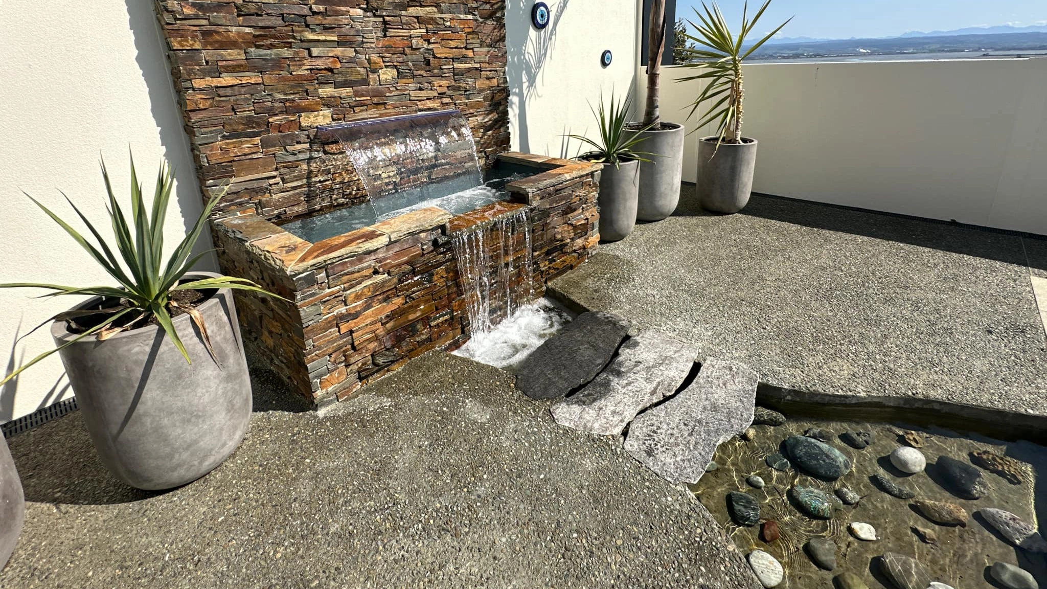 The Reflection with Auto Cover 9.90m fibreglass swimming pool is installed in a metropolitan region of Stoke, New Zealand. The house is surrounded by grey tiles, a small waterfall and natural greenery with mountains in the background. The pool colour is Crystal Blue, which stands out from the tranquil landscape and white house.