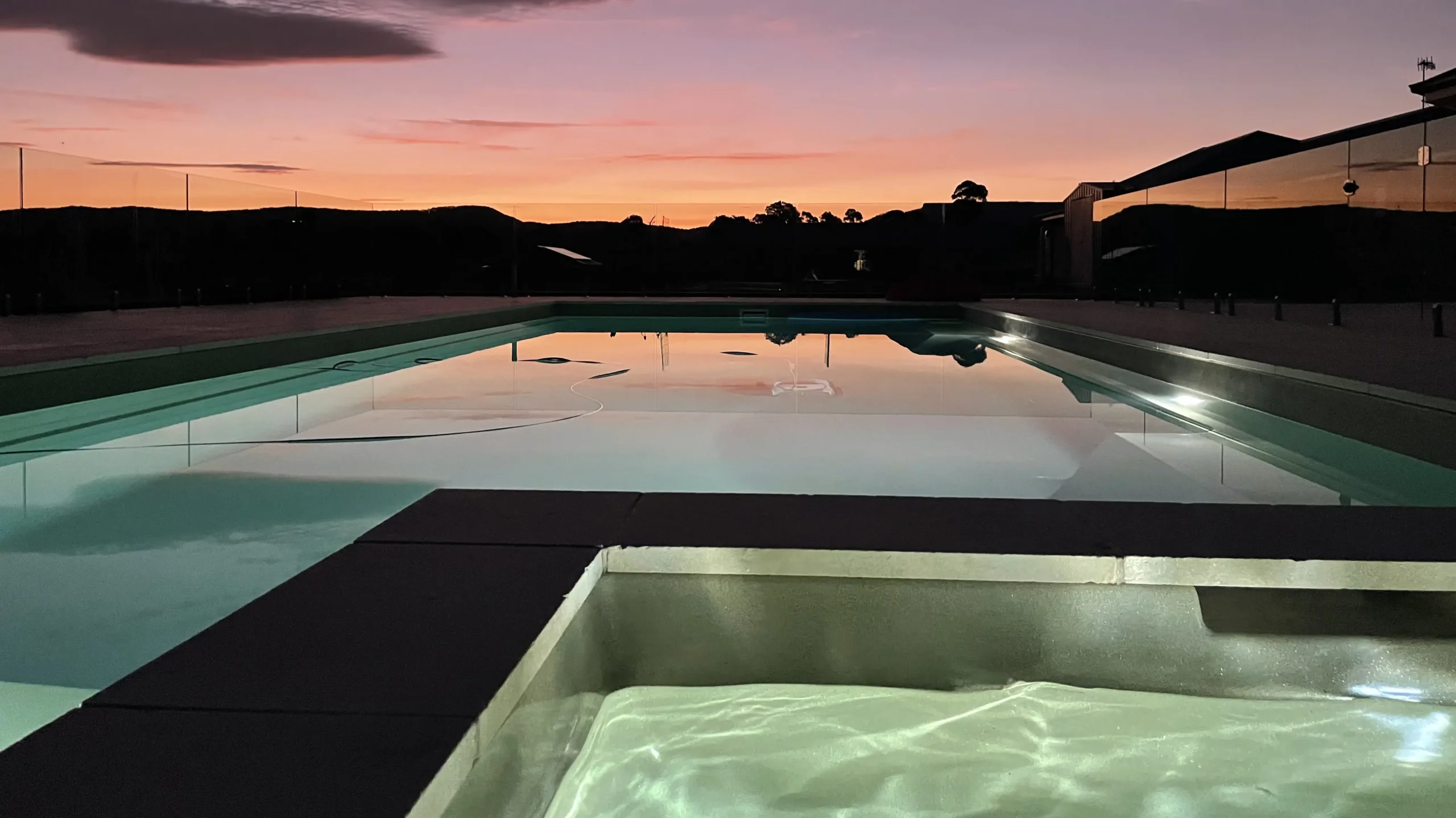The Absolute 12.00m fibreglass swimming pool is in the middle of the backyard, with white tiles and greenery surrounding it. The pool colour is Diamond Sand, which makes the fibreglass swimming pool stand out from the modern countryside house and the dawn orange sky.